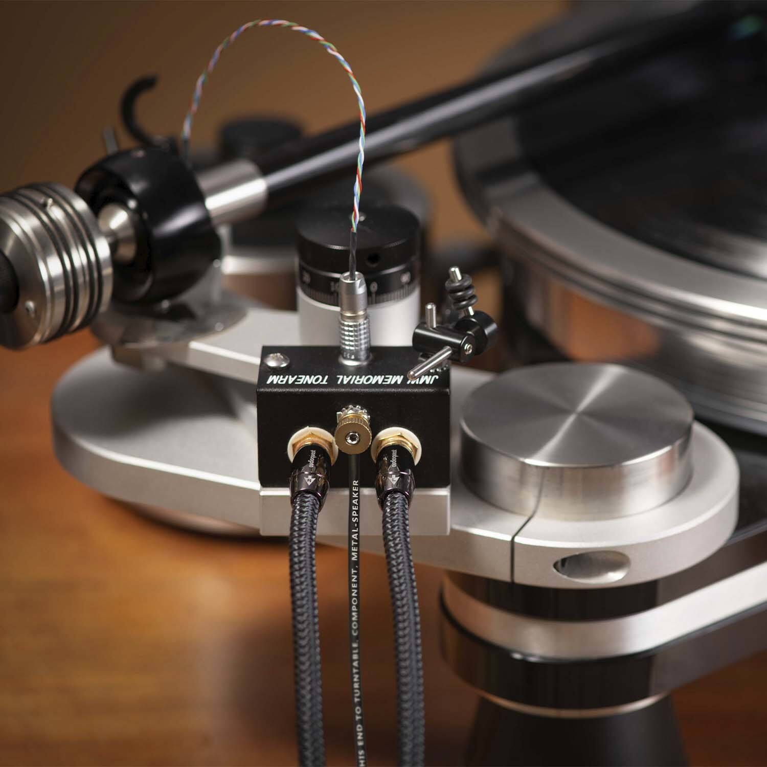 The image shows a turntable with a tonearm and a speaker component. The tonearm is connected to the turntable, and there is a metal-speaker component present. The turntable is placed on a wooden table.