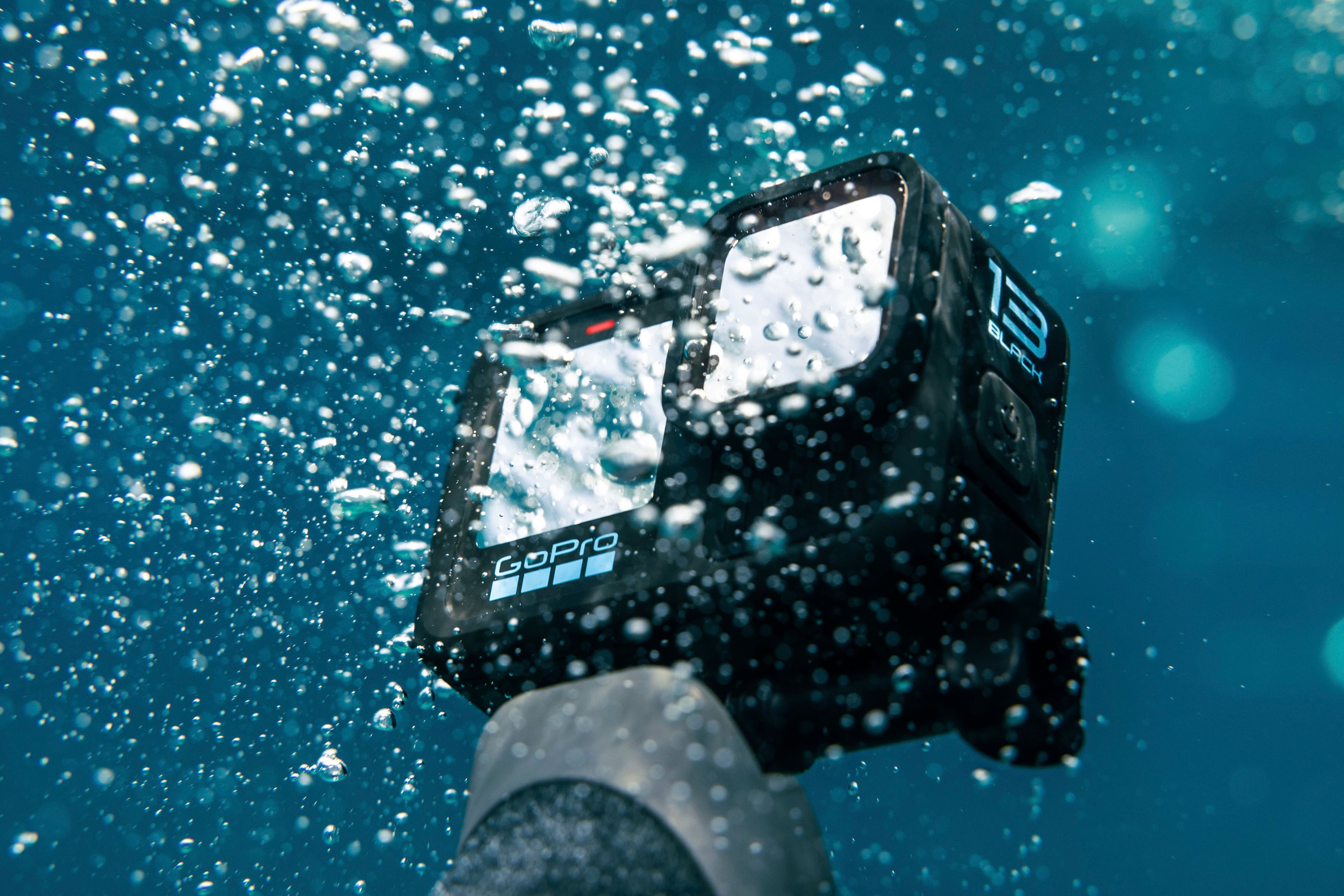 The image shows a person holding a black GoPro camera underwater, capturing the beauty of the ocean. The camera is positioned in front of the person, and the reflection of the person can be seen in the camera lens. The scene is bathed in bubbles, adding to the underwater atmosphere. The person is likely enjoying their time snorkeling or diving, taking advantage of the clear water and the opportunity to capture stunning underwater footage.