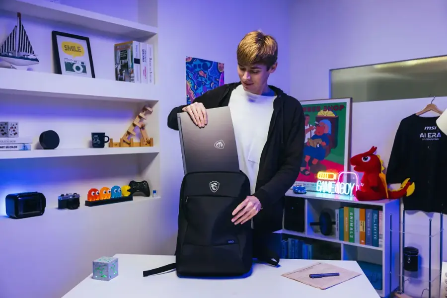 The image shows a man standing in front of a table, holding a laptop bag. He is likely preparing to put the laptop into the bag. The table is surrounded by various items, including several books, a cup, and a backpack. The man is wearing a black jacket, and there is a teddy bear nearby. The scene suggests that the man might be a student or a professional who is organizing his belongings or preparing for a trip.