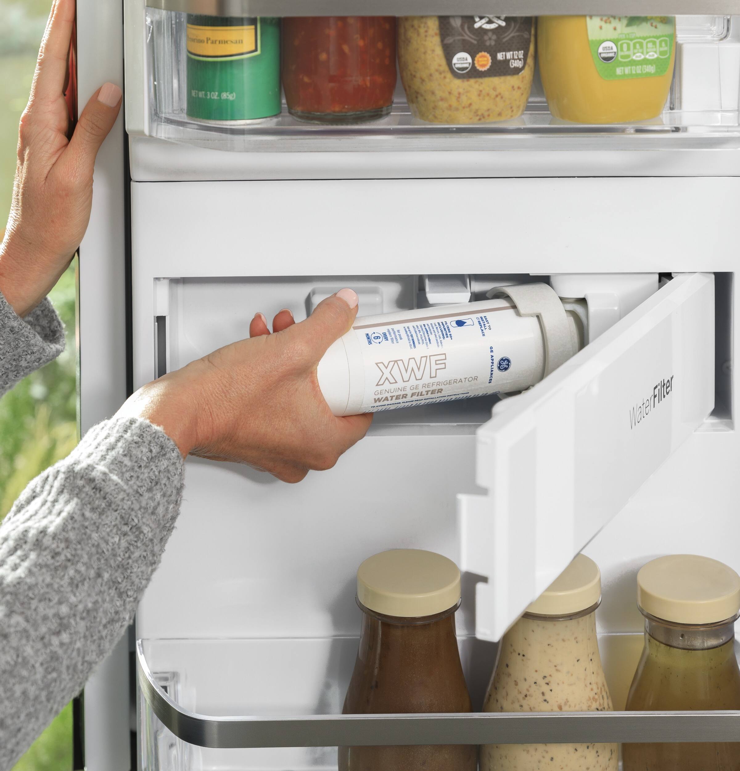 The image shows a person opening a refrigerator door and pulling out a bottle of water. The refrigerator is filled with various bottles, including some containing sauces and juices. The person is reaching for a bottle of water, which is placed on the door shelf. The refrigerator is well-stocked with a variety of items, making it a convenient place to store and access food and beverages.