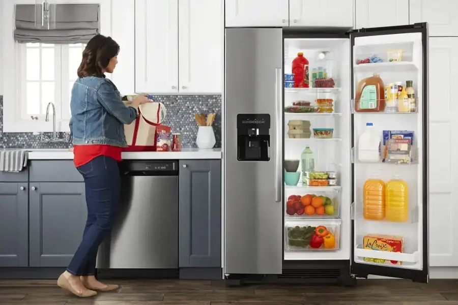 The image shows a woman standing in a kitchen, opening a refrigerator. She is wearing a red shirt and jeans. The refrigerator is filled with various food items, including multiple bottles, oranges, and a bowl. The woman is holding a handbag, and there is a sink and a dining table in the kitchen. The scene suggests that the woman is preparing a meal or looking for something to eat or drink.