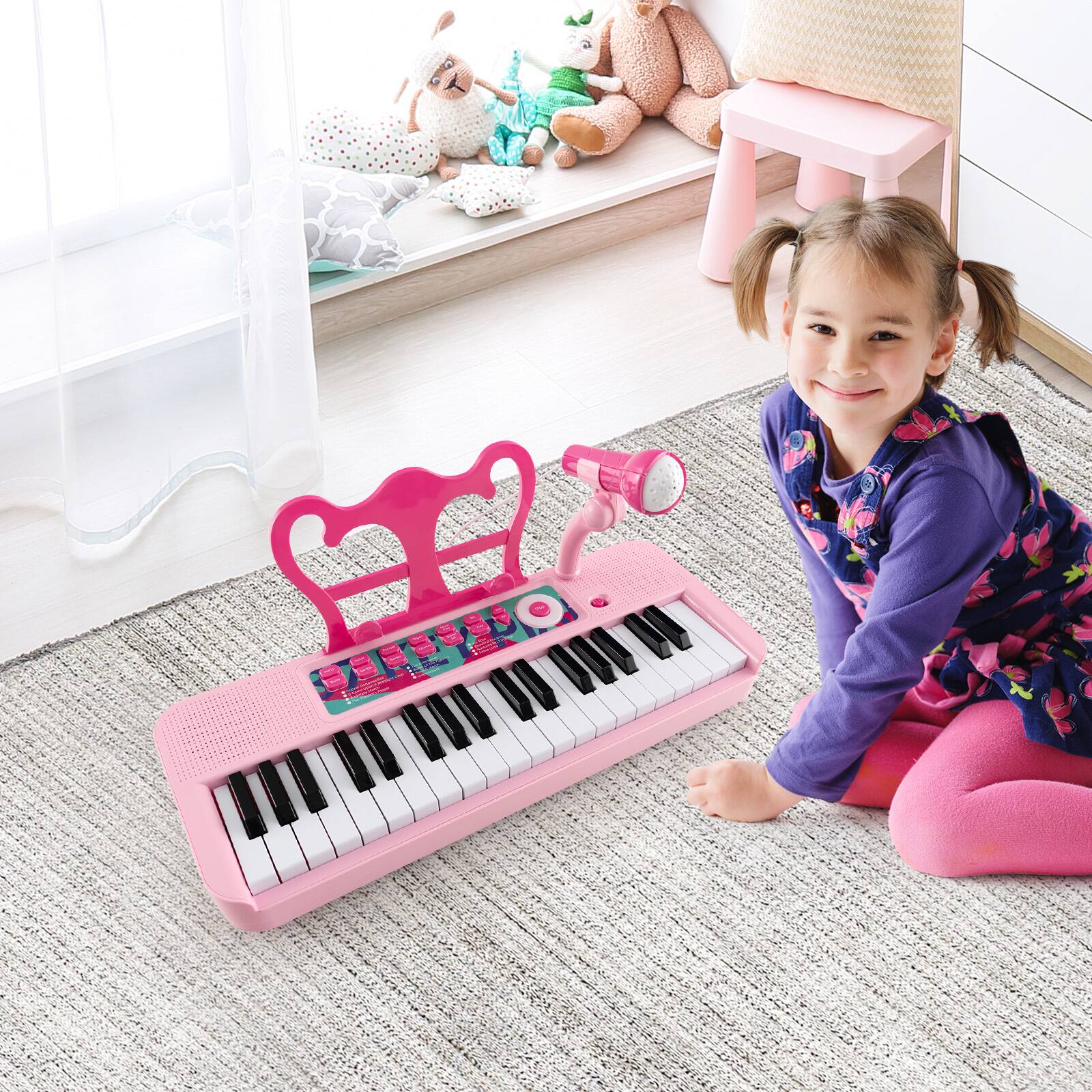The image shows a young girl sitting on the floor in front of a pink keyboard. She is smiling and appears to be enjoying herself. The scene also includes a teddy bear and a chair nearby.