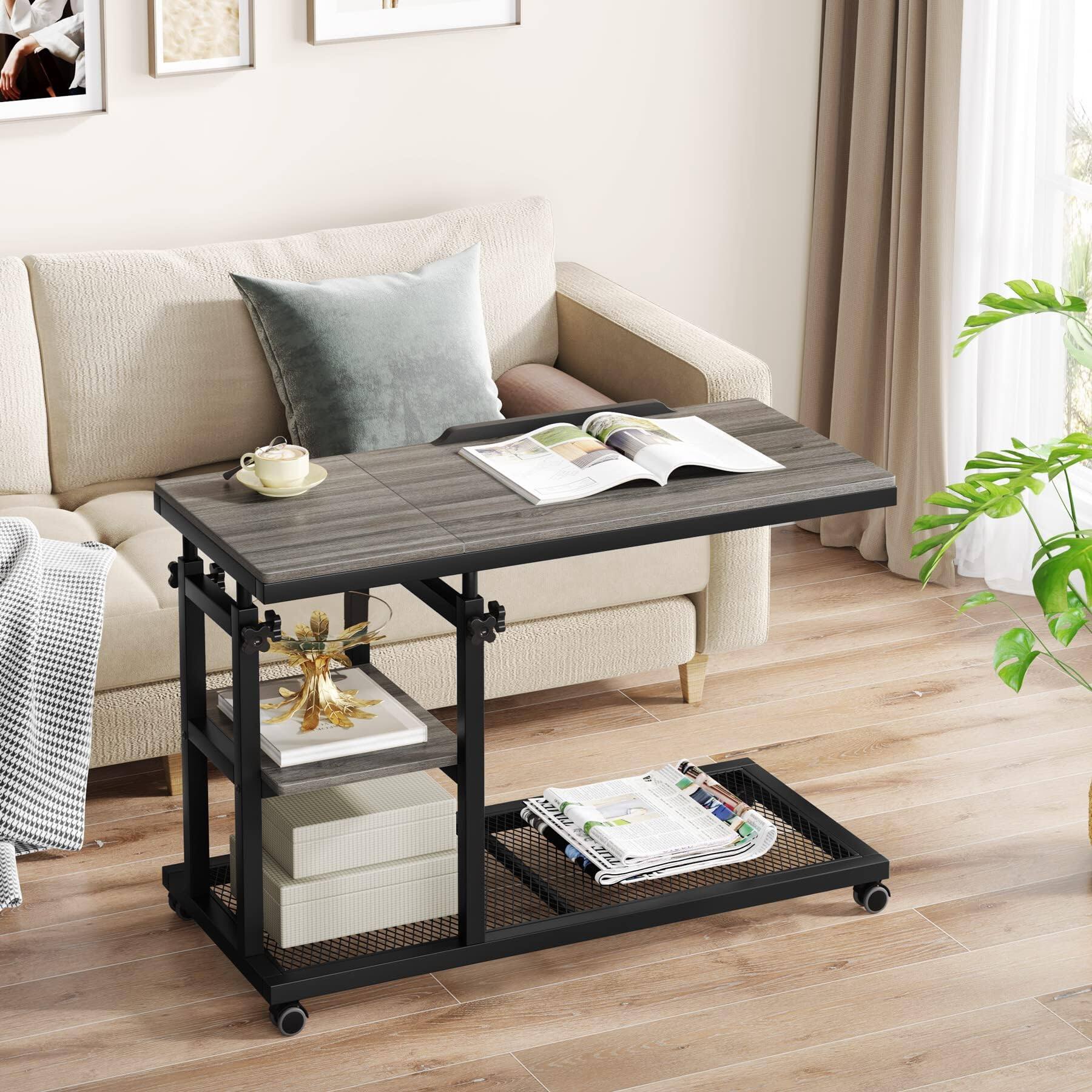 The image shows a modern living room setting featuring a beige sofa with a gray cushion. In front of the sofa is a black metal side table with a wooden top. On the table, there is a white teacup and saucer, and an open magazine. Below the table, there are two shelves: the top shelf holds a decorative gold and white box, and the bottom shelf contains a stack of books and a folded newspaper. The room has light wooden flooring, a large window with beige curtains, and a green plant in the corner.