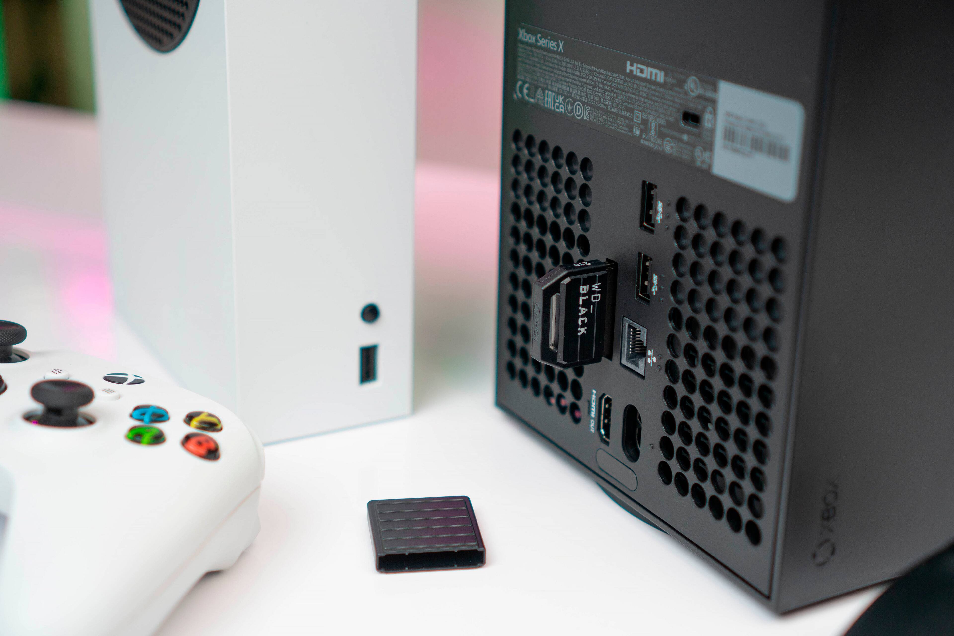 The image shows a black Xbox Series X gaming console sitting next to a white Xbox Series X. The black console has a sticker on it, and there is a small black box nearby. The scene also includes a video game controller, possibly for the Xbox.
