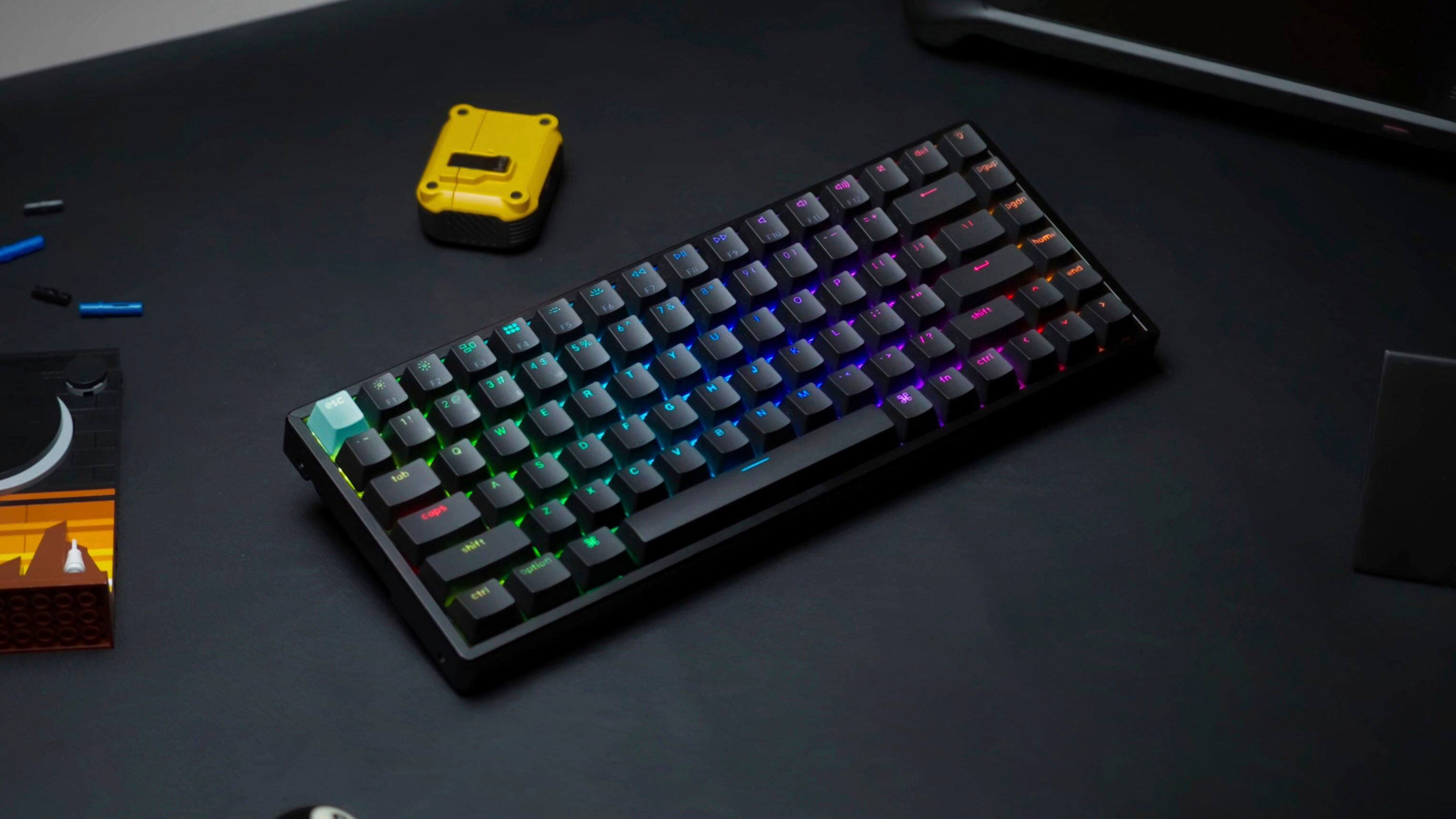 The image shows a colorful keyboard sitting on a black table. The keyboard has a unique design, featuring a combination of letters and numbers. The table also has a toy car and a remote control nearby.