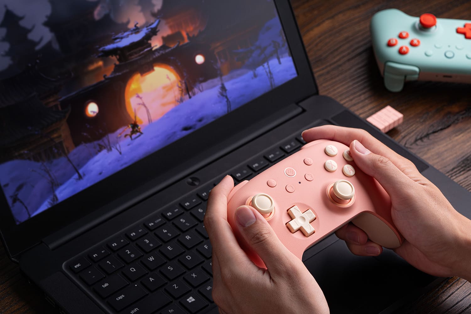 A person is using a laptop with a pink game controller. They are playing a game on the laptop, possibly using the game controller to control the game. The game controller is placed on the laptop keyboard, and the person's hands are on the controller.