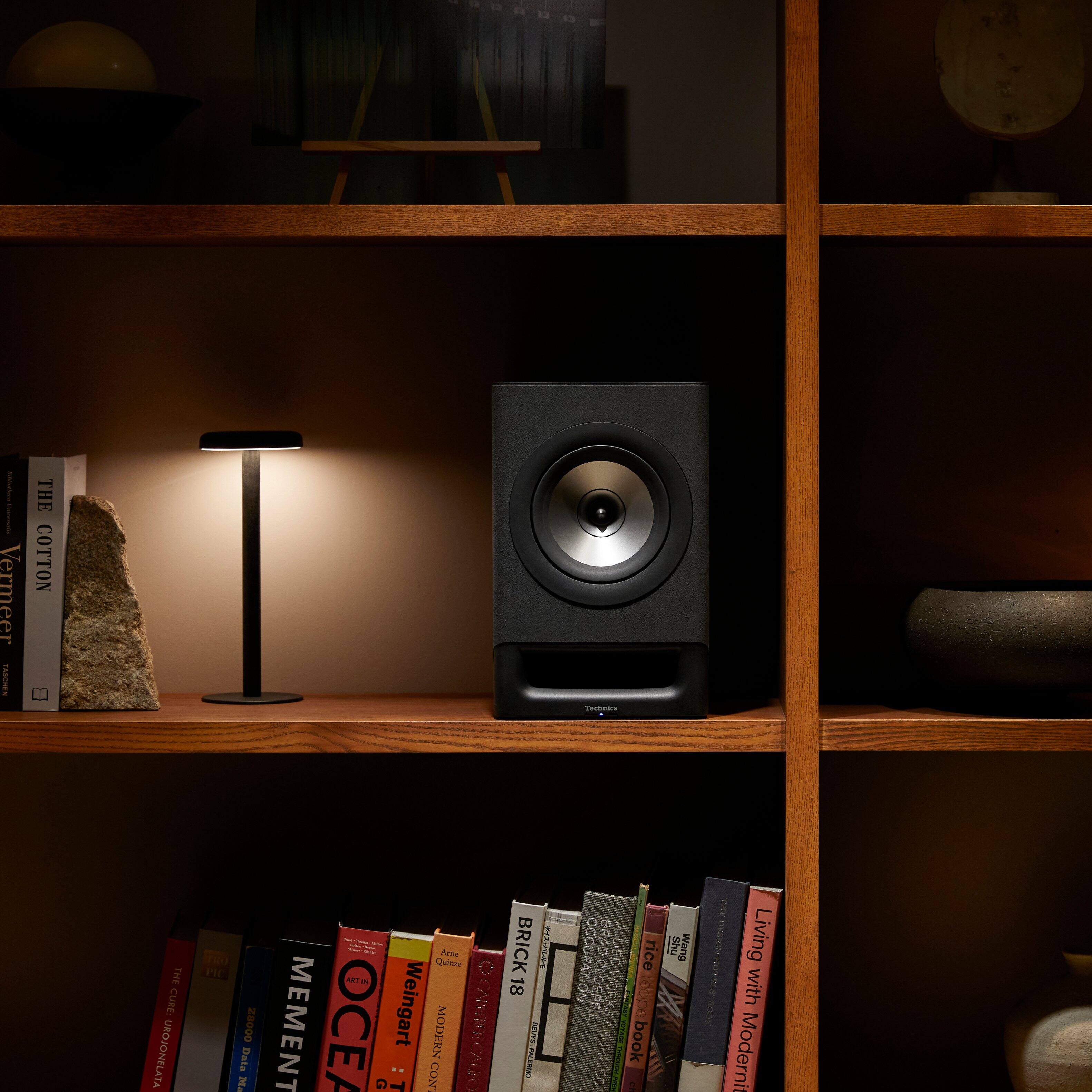 The image features a bookshelf with various books and a speaker. The books are arranged in different sections, with some books placed on the left side of the shelf, some on the right side, and others in the middle. The speaker is positioned in the middle of the shelf, surrounded by the books. The bookshelf is filled with a variety of books, creating an organized and visually appealing display.