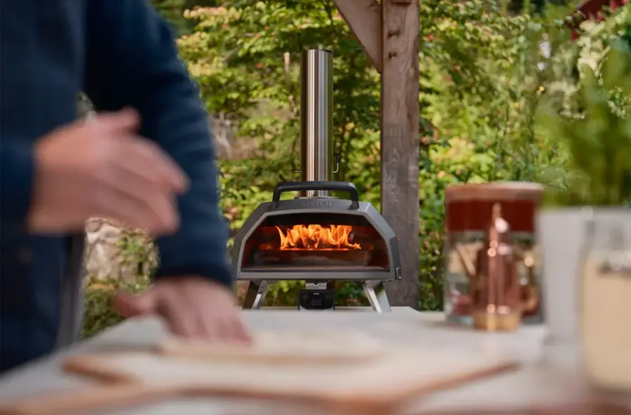 The image shows a man standing near a small pizza oven, which is located on a table. The oven is lit, indicating that it is being used to cook pizza. The man appears to be preparing the pizza, possibly adding toppings or adjusting the cooking process. The scene takes place outdoors, with a dining table nearby. There are also several potted plants in the area, adding a touch of greenery to the setting.