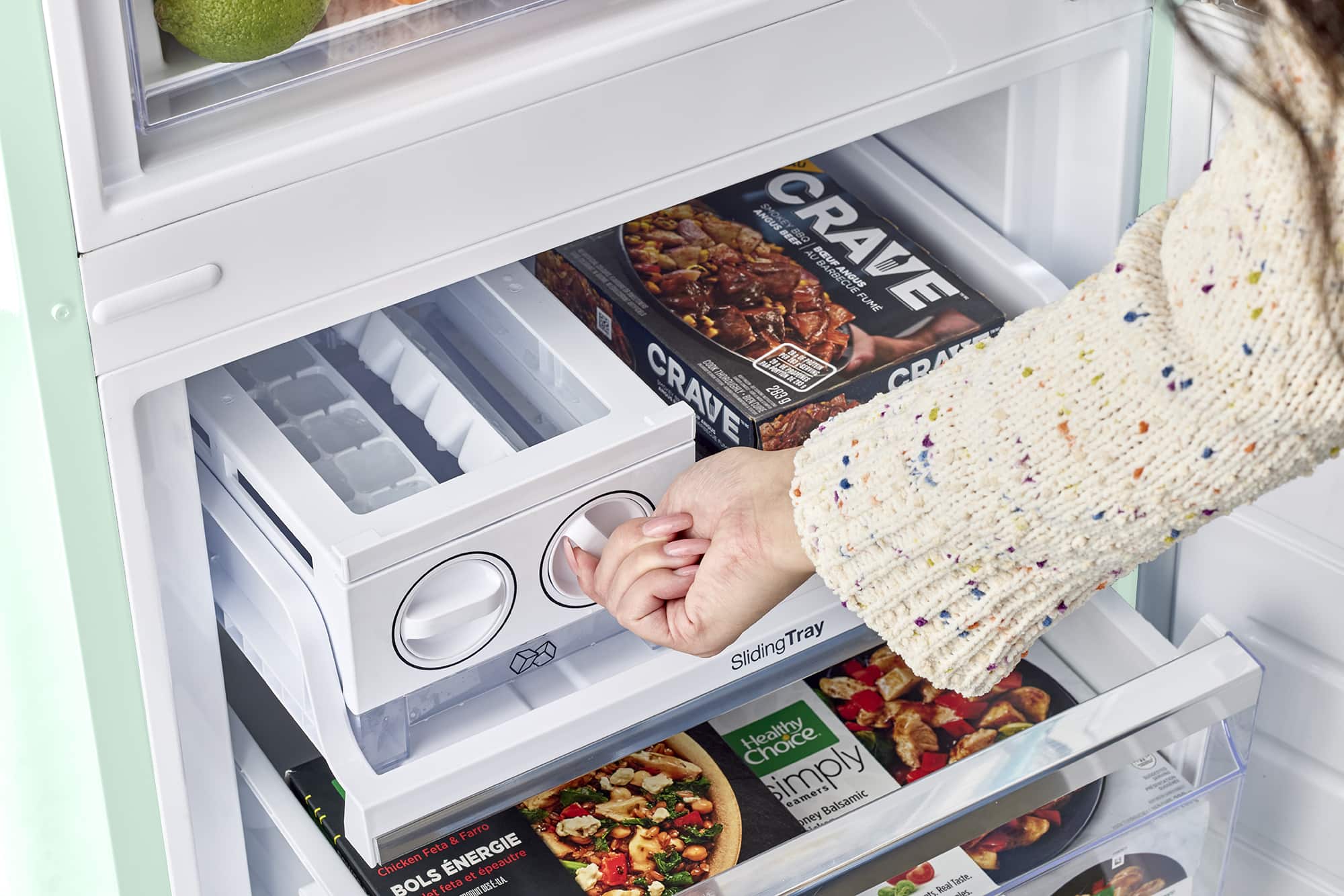 A person is opening a refrigerator, which contains a variety of food items. The refrigerator has a sliding drawer with a tray of food, including a pizza, and a drawer with a Crave beef product. There are also apples and oranges stored in the refrigerator.