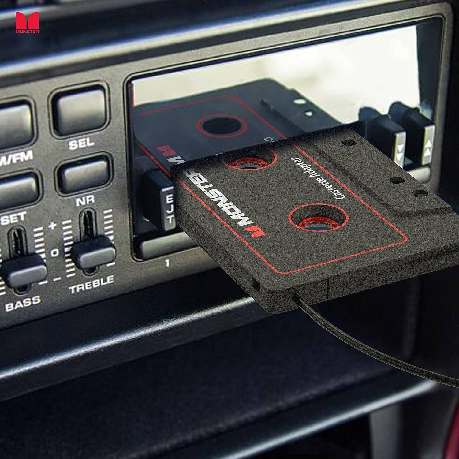 The text on the image reads: "MUNSTER didk SEL A/FM NR SET + O : - - E TREBLE BASS C Adapter tte MONSTER Cassette JT 1". This is a description of a cassette adapter that is being used in a car. The adapter is designed to connect a cassette tape player to the car's sound system, allowing the user to play cassette tapes through the car's speakers. The adapter is made by the company Monster and is compatible with a variety of car models.
