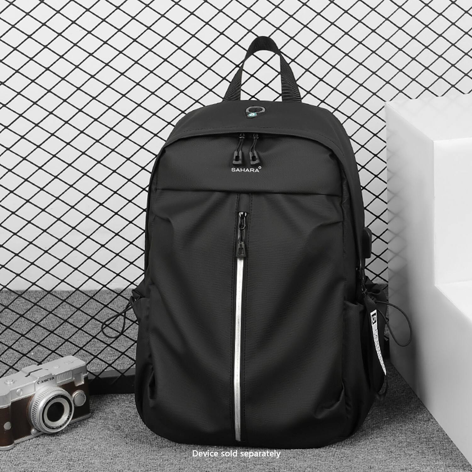 The image features a black backpack and a camera. The backpack is a Sahara brand, and it is sitting on the floor next to the camera. The text on the image reads "Device sold separately."
