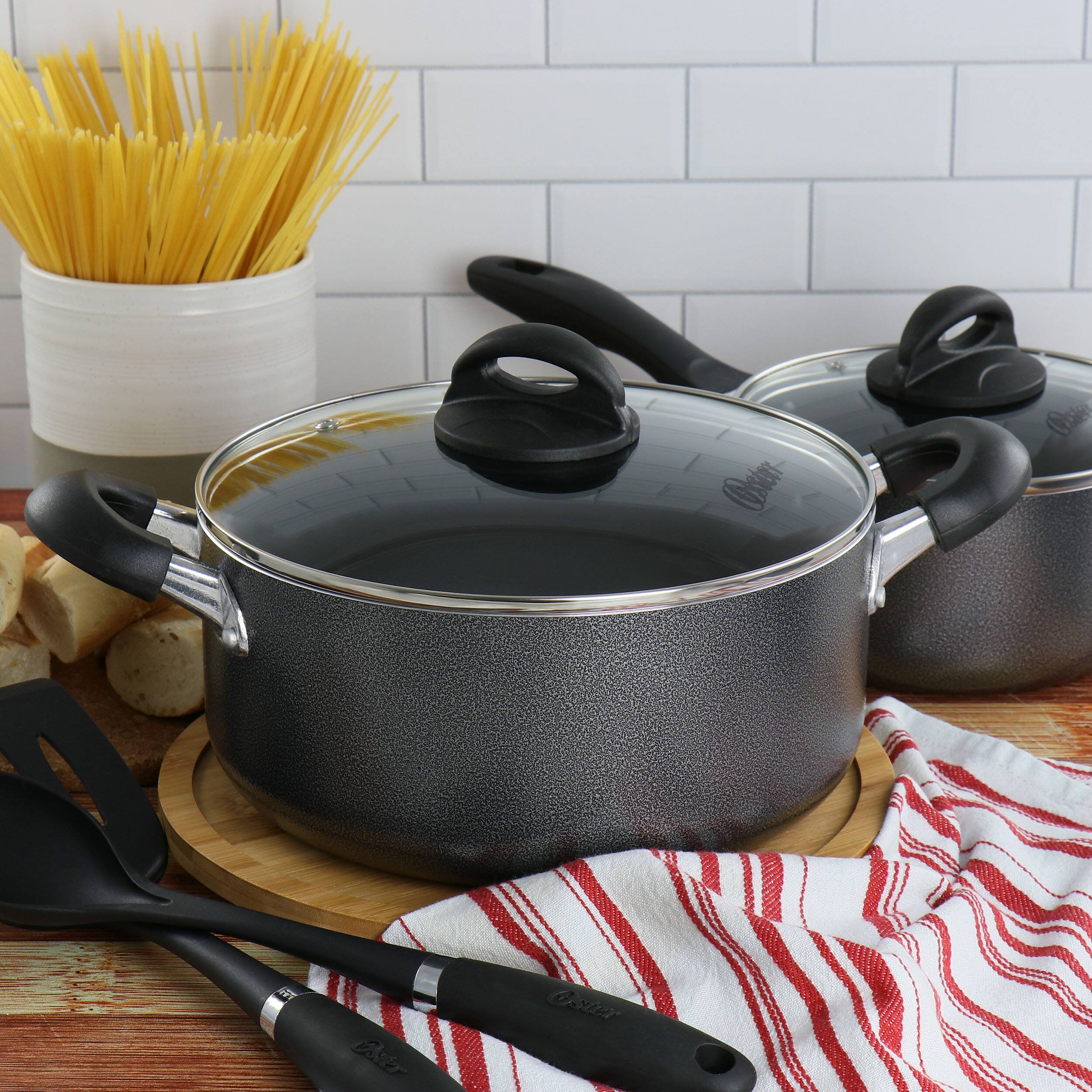 The image features a table with two large pots, one of which is a silver pot with a black handle. There are also two black pots on the table, along with a spoon and a fork. The table also has a bowl of pasta and a basket of pasta. The scene seems to be set in a kitchen, with a tiled wall in the background.