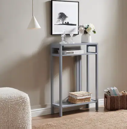 In the middle of the room, there is a small, light blue side table with a white lamp hanging above it. On the table, there are two clear glasses, a white vase with flowers, and a book titled "EXPLORE THE WORLD." Below the table, there is a woven basket. To the right, there is a wicker basket with some books and a wooden object.