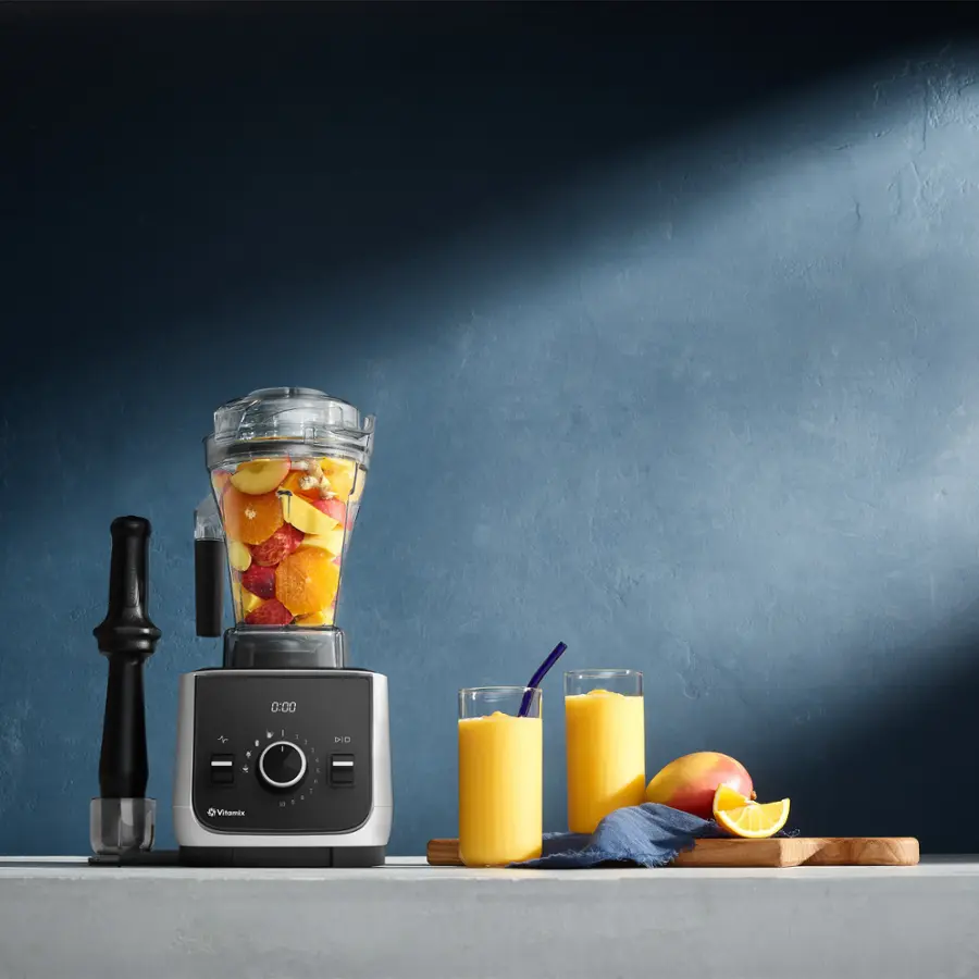 The image shows a blender filled with fruit, such as apples and oranges, and a pitcher of juice. The blender is placed on a counter, and there is a cutting board nearby. The scene is set against a blue wall, and a knife is also visible in the image.