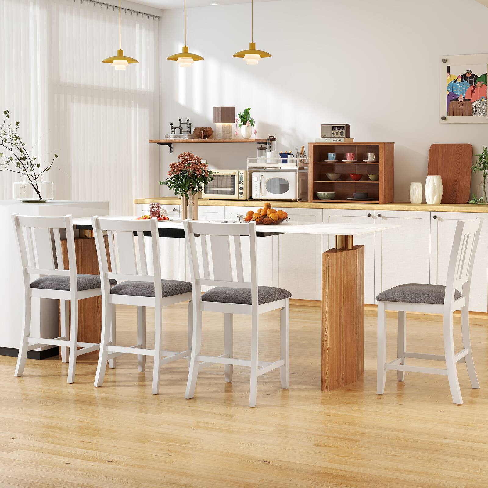 The image shows a modern kitchen with a dining area. The kitchen features white cabinets, a wooden countertop, and a light wooden floor. There are four white chairs with gray cushions around a wooden table. The table has a vase with flowers and a bowl of oranges. Above the table, there are three pendant lights with a gold finish. The kitchen is equipped with a microwave, a toaster, and various kitchenware on the shelves. A painting hangs on the wall, and there are plants and a window with blinds.