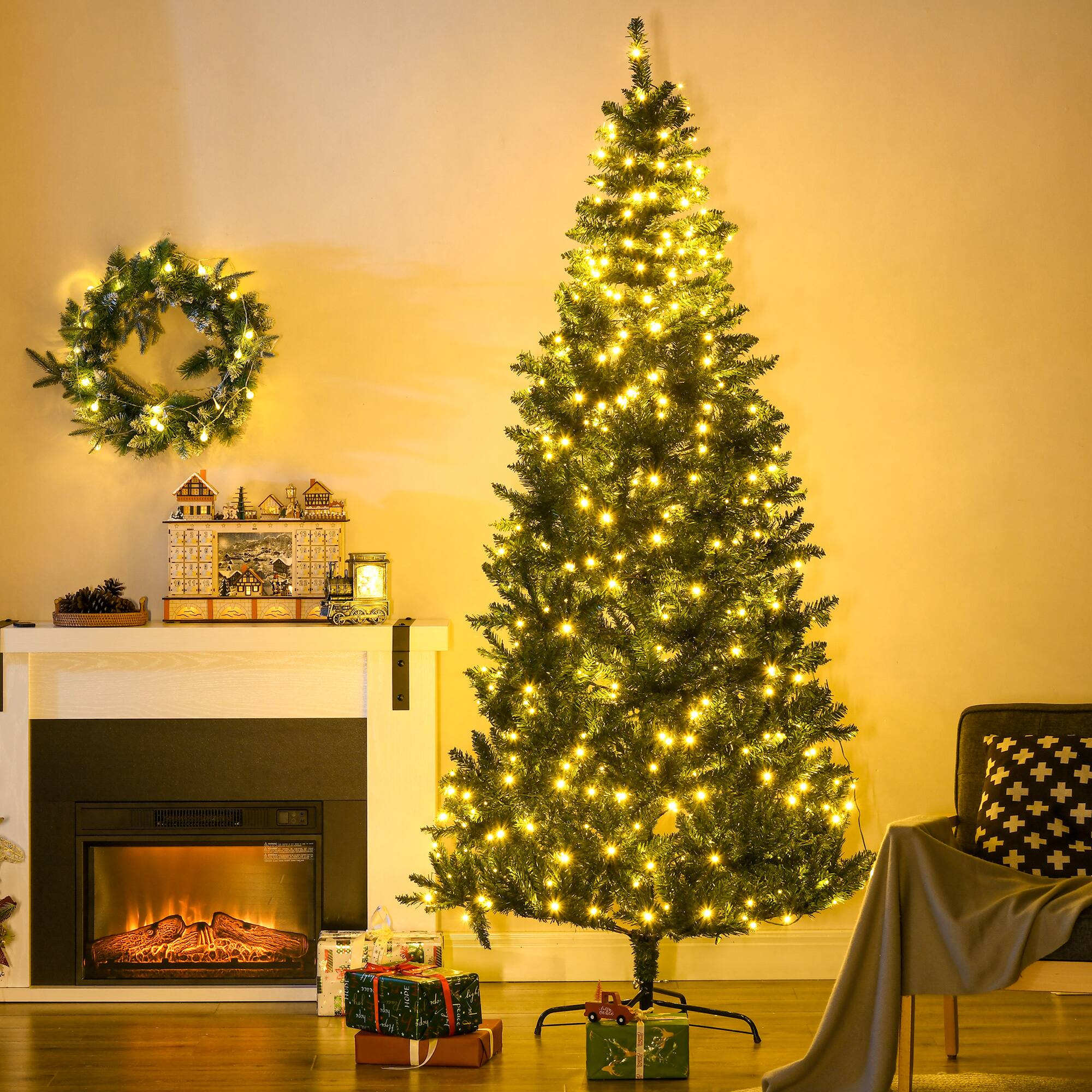 The image shows a cozy, festive scene featuring a decorated Christmas tree with warm yellow lights. The tree is placed next to a fireplace, which has a lit electric firebox. Above the fireplace, there is a wreath with lights and a mantel decorated with small Christmas-themed items. Several wrapped presents are placed under the tree and near the fireplace. The room has a warm, inviting ambiance with soft lighting.