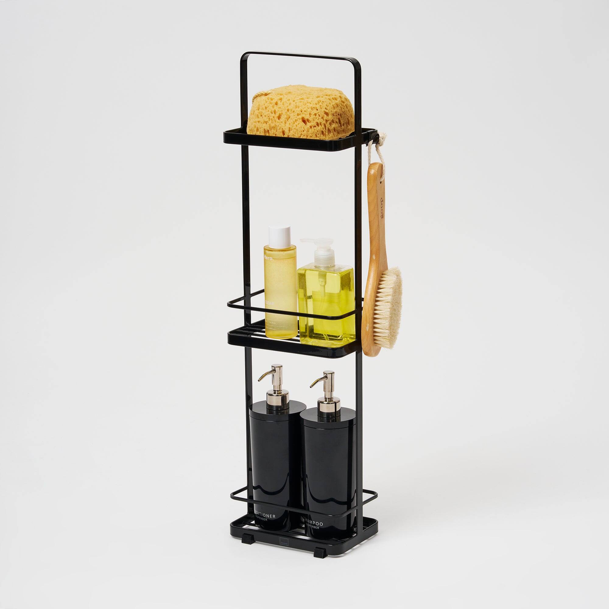 There is a black metal rack with three shelves. On the top shelf, there is a large, textured soap bar. The middle shelf holds two bottles of liquid soap, one yellow and one clear, with white pump dispensers. On the right side of the middle shelf, there is a wooden brush with a yellow handle. The bottom shelf has two black soap dispensers with gold-colored pumps. The rack is labeled "BATHROOM" on the front.