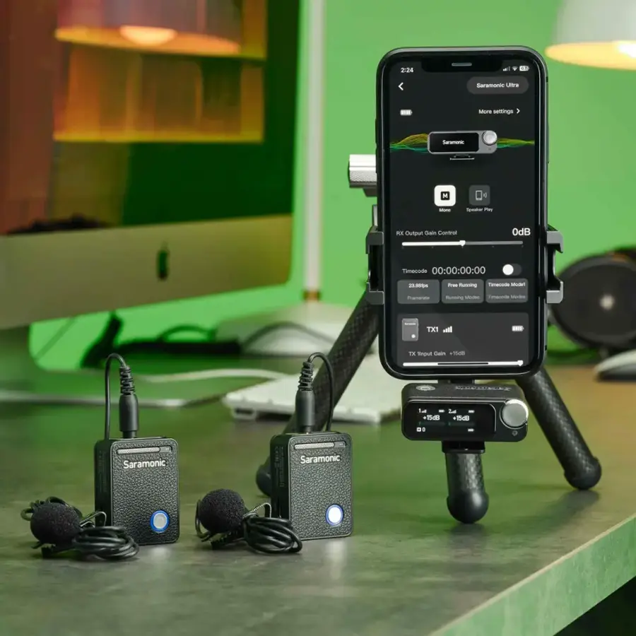 The image features a cell phone propped up on a stand, with a microphone attached to it. The cell phone is displaying a screen with various settings, including timecode, gain control, and other audio-related options. The stand is positioned on a desk, and there is a computer monitor nearby. Additionally, there are two microphones in the scene, one of which is connected to the cell phone, and the other is placed separately.