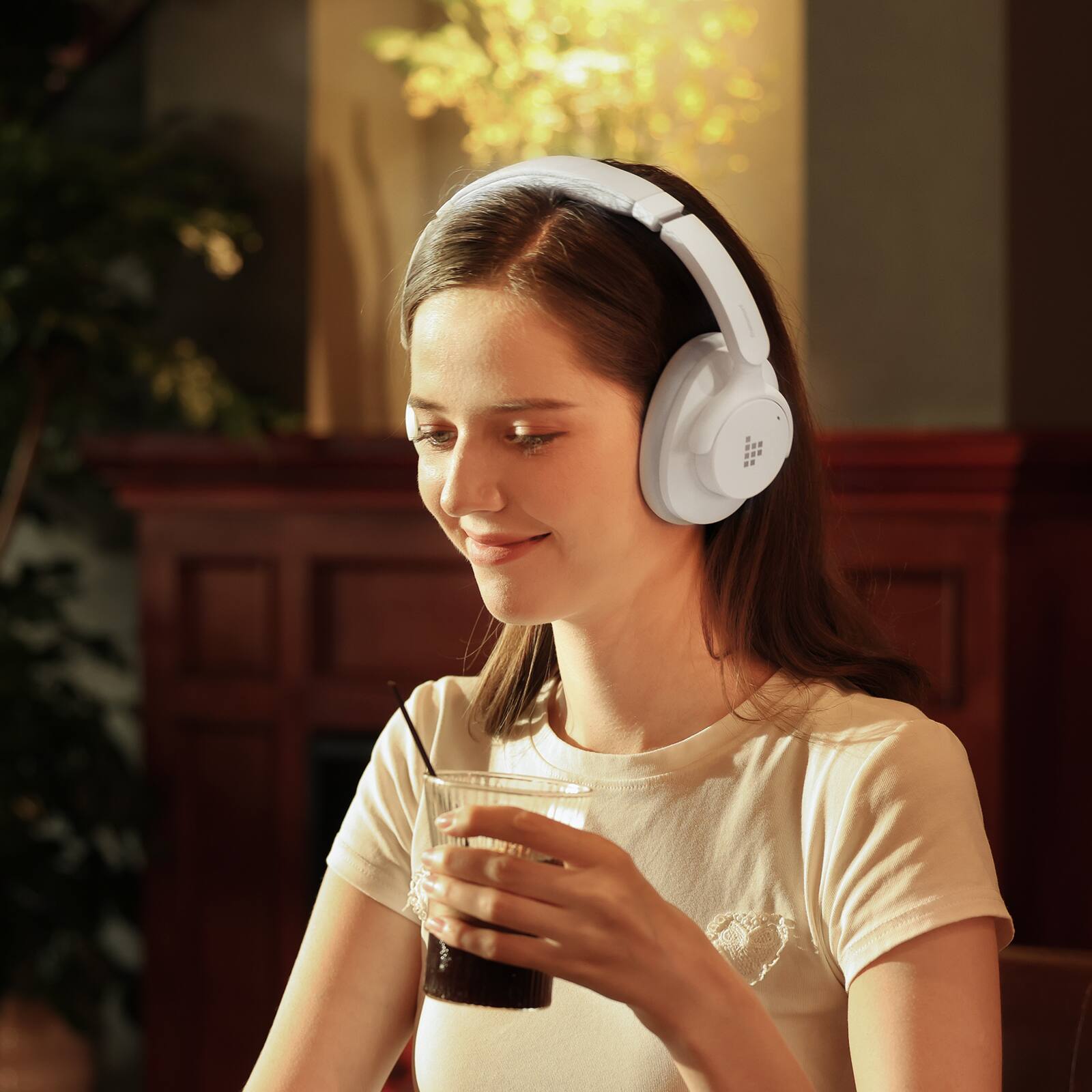 The image shows a woman wearing headphones and holding a drink, possibly a cup of coffee or a glass of soda. She is smiling and appears to be enjoying her beverage. The woman is standing in a room, and there is a potted plant nearby. The scene captures a moment of relaxation and leisure for the woman as she listens to music or an audio program through her headphones.