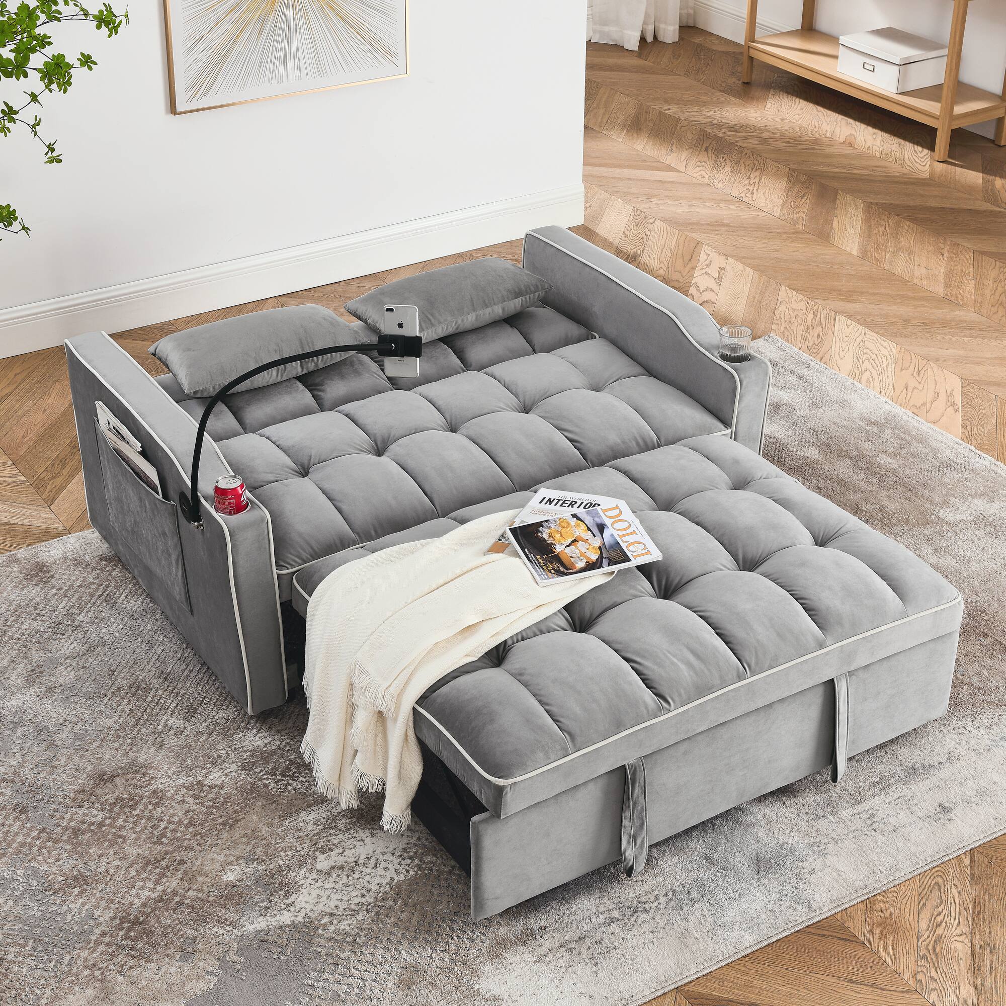 The image shows a modern, tufted gray sofa with a matching chaise lounge. The sofa is placed on a textured rug, and there is a white throw blanket draped over it. A newspaper and a can of soda are on the sofa. The room has wooden herringbone flooring and a piece of abstract art on the wall.