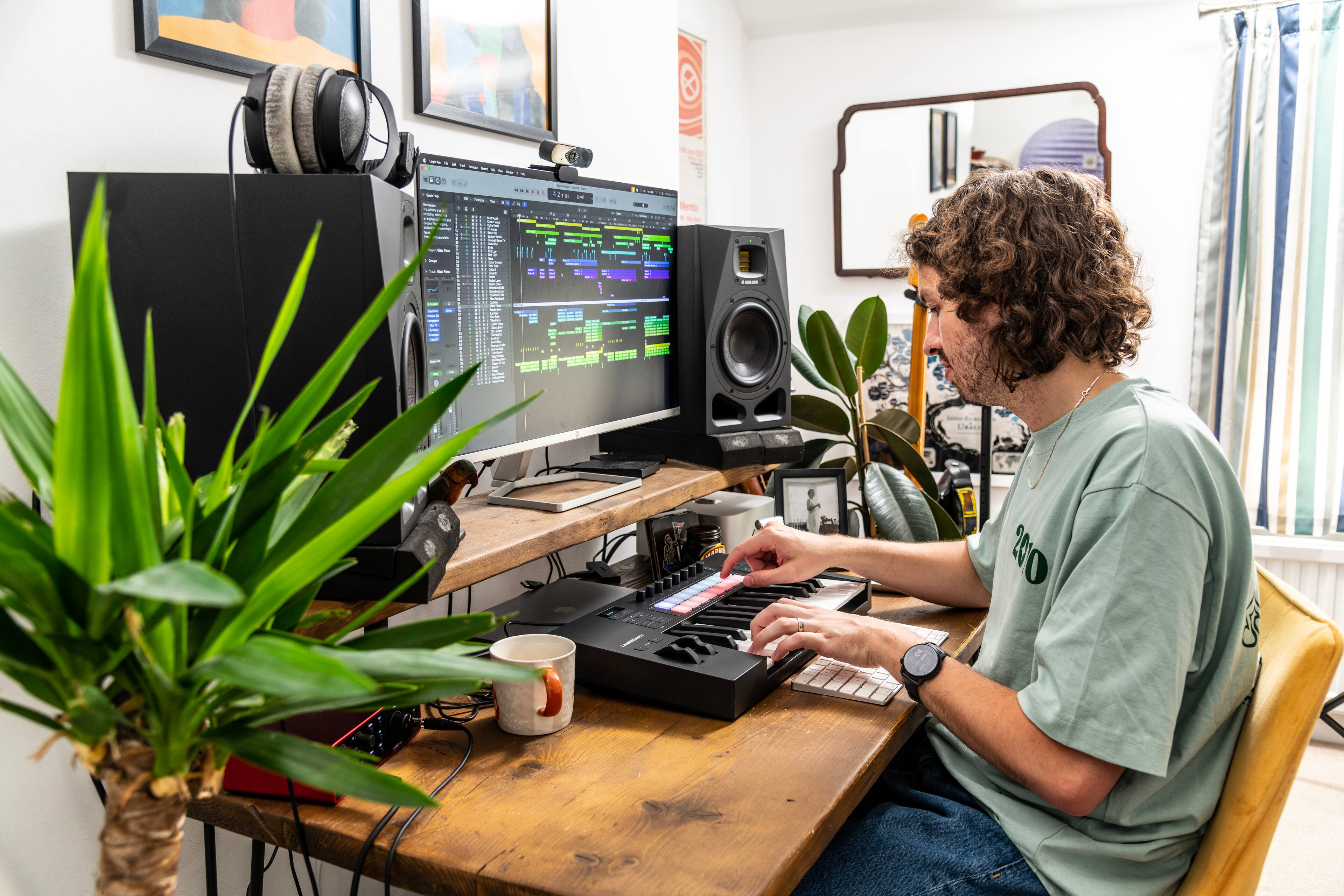The man is sitting at a desk with a computer, keyboard, and mouse. He is wearing a green shirt and has a beard. There is a potted plant next to him, and a cup is placed on the desk. The man is likely working or using the computer for some purpose.