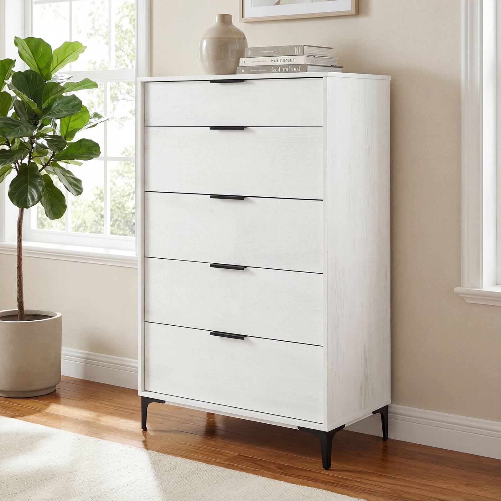 This is a white, five-drawer storage unit with black handles. It has a light wood finish and is placed against a wall near a window. There is a potted plant to the left of the unit, and a vase and books on top of it.