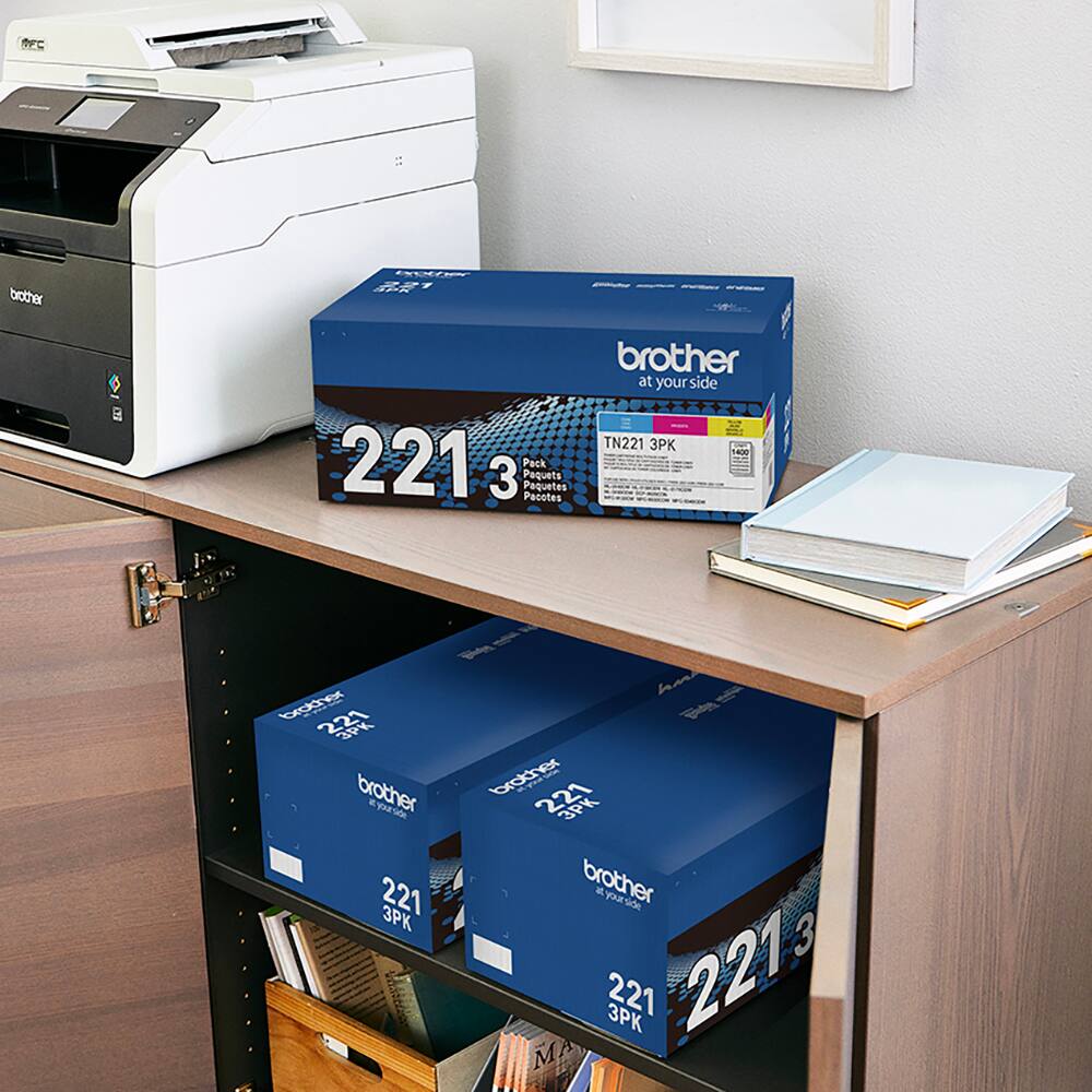 The image features a desk with a printer and a box of Brother toner cartridges. The box contains three toner cartridges, each with a capacity of 2213 pages. The cartridges are designed for use with Brother printers, providing reliable and high-quality printing. The desk also has a book and a notebook, suggesting that it is a workspace for various tasks and projects.