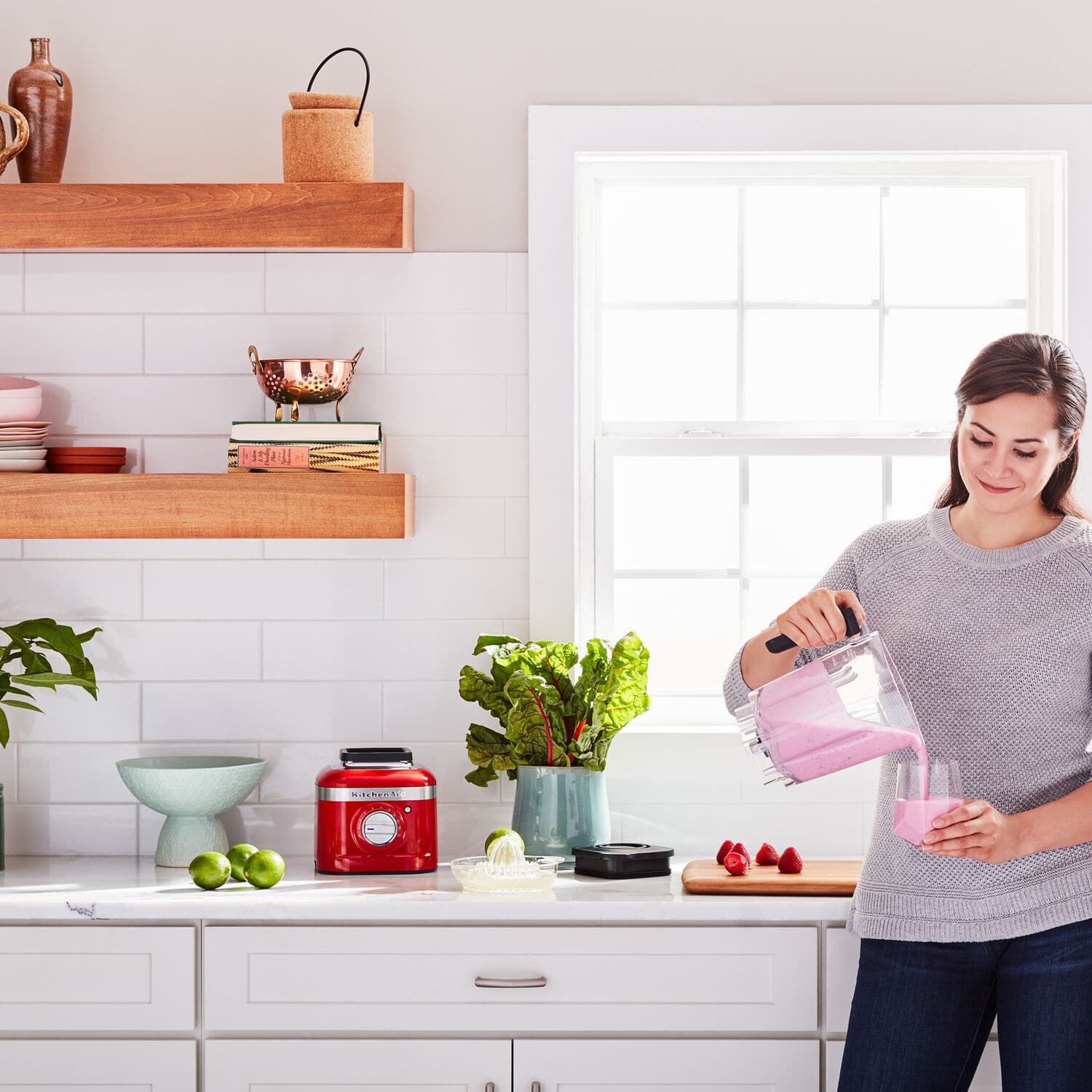 The image shows a woman standing in a kitchen, preparing a smoothie in a pink blender. She is pouring the smoothie into a cup. The kitchen is well-equipped with various items, including a sink, a bowl, a potted plant, and a vase. There are also several books scattered around the kitchen, possibly cookbooks or reference materials for the woman's cooking endeavors.