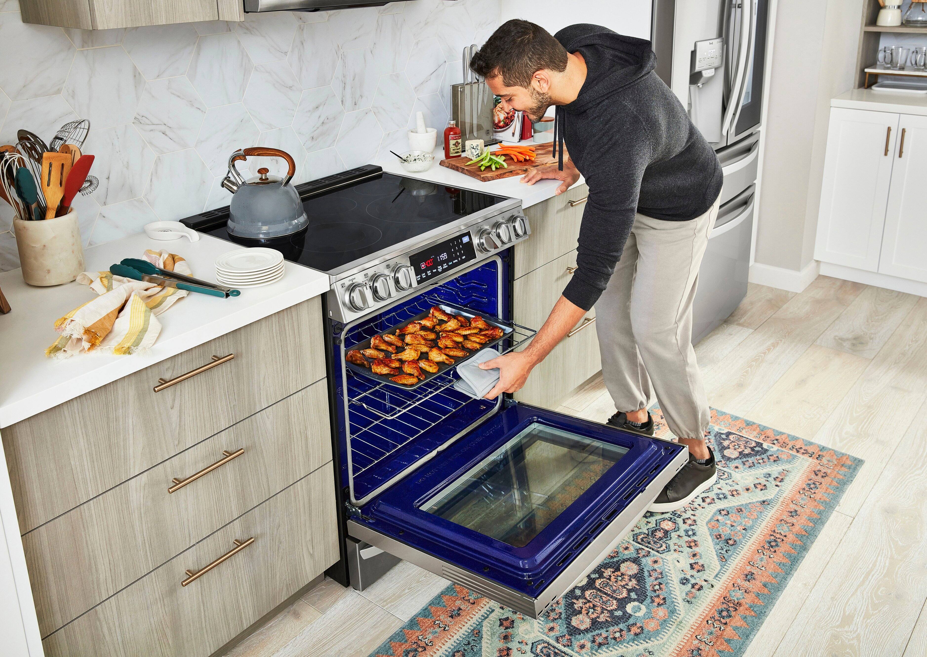 The man is standing in a kitchen, wearing a black sweater and holding a tray of food out of the oven. The oven is blue and has a glass door. The man is also holding a spray bottle, possibly for cleaning or cooking purposes. The kitchen is well-equipped with various utensils, including a knife, a spoon, a fork, and a bowl. There is also a sink and a microwave in the kitchen.
