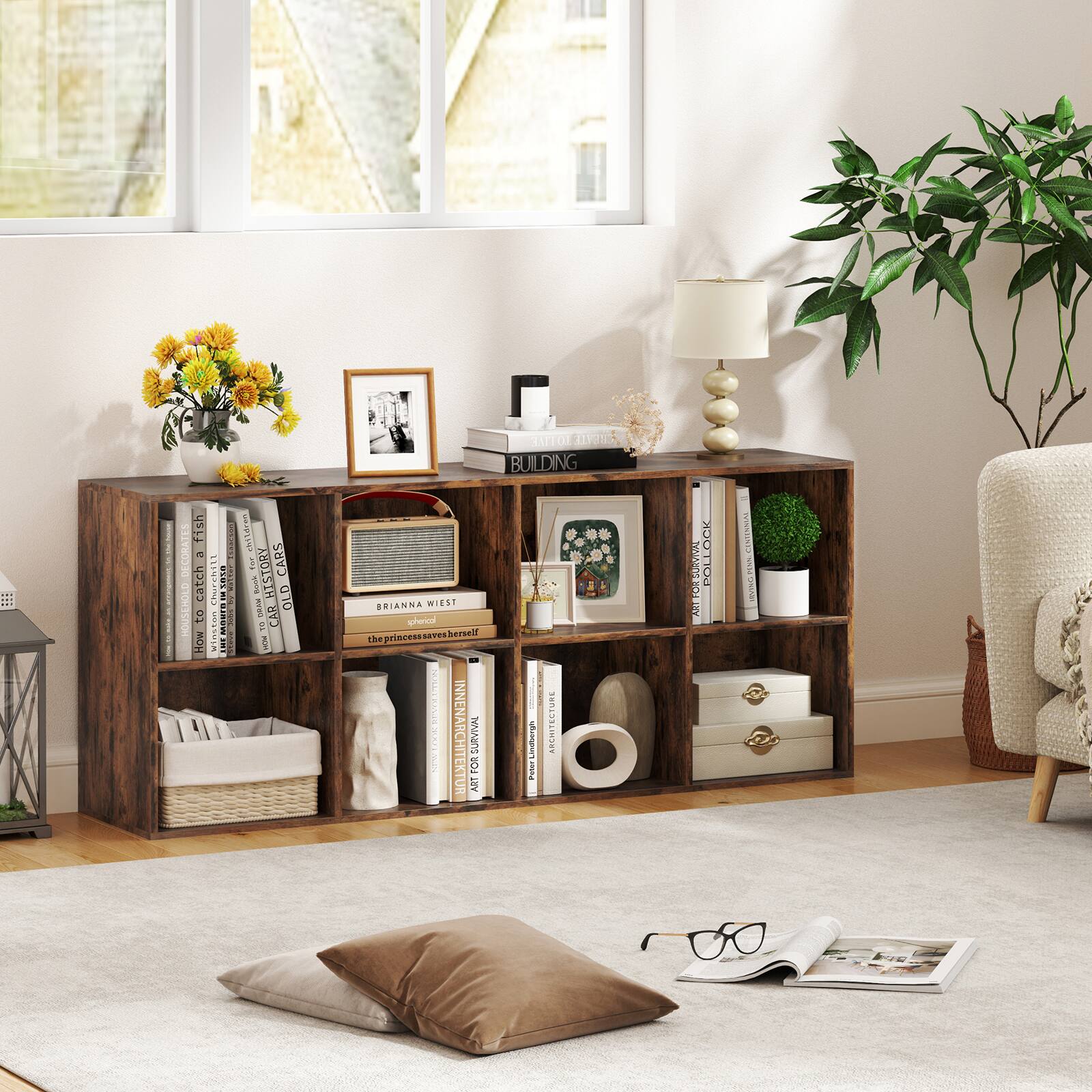 The image shows a living room with a bookshelf filled with books, a lamp, and a potted plant. There is also a chair and a couch in the room. The bookshelf is located next to a window, and there are several books of different sizes on it. Additionally, there is a remote control placed on the couch, and a pair of glasses is resting on the couch. The room has a cozy and inviting atmosphere, perfect for relaxation and reading.