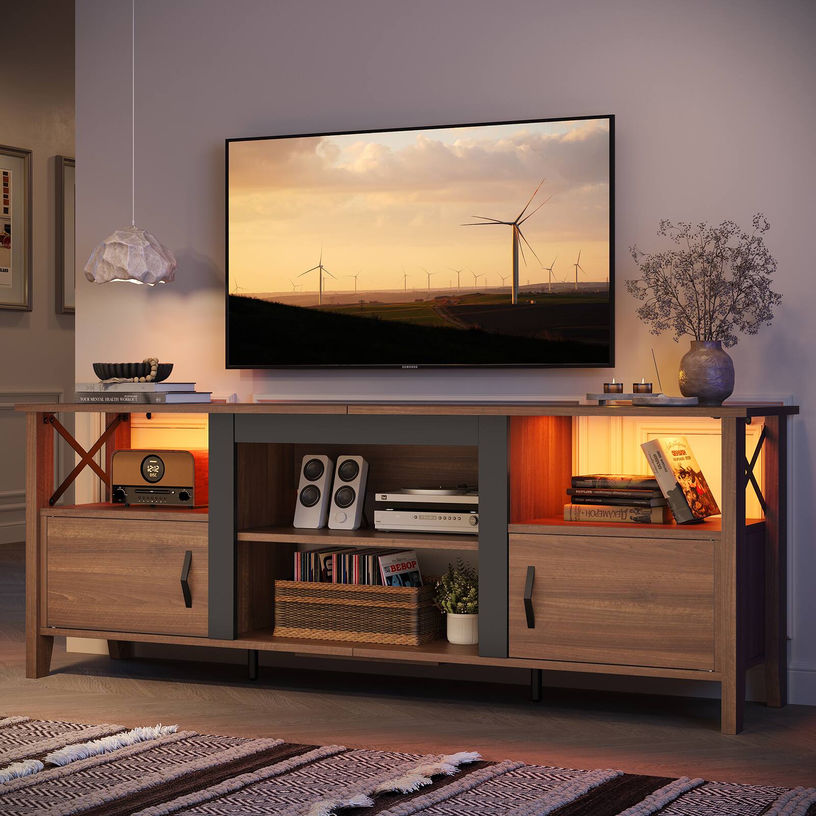 The image features a living room with a large flat screen TV mounted on the wall. The TV is displaying a picture of windmills. The room also has a wooden entertainment center with a speaker and a clock. There are several books scattered around the room, and a potted plant is placed nearby. Additionally, there is a vase and a bowl in the room, adding to the overall decor.