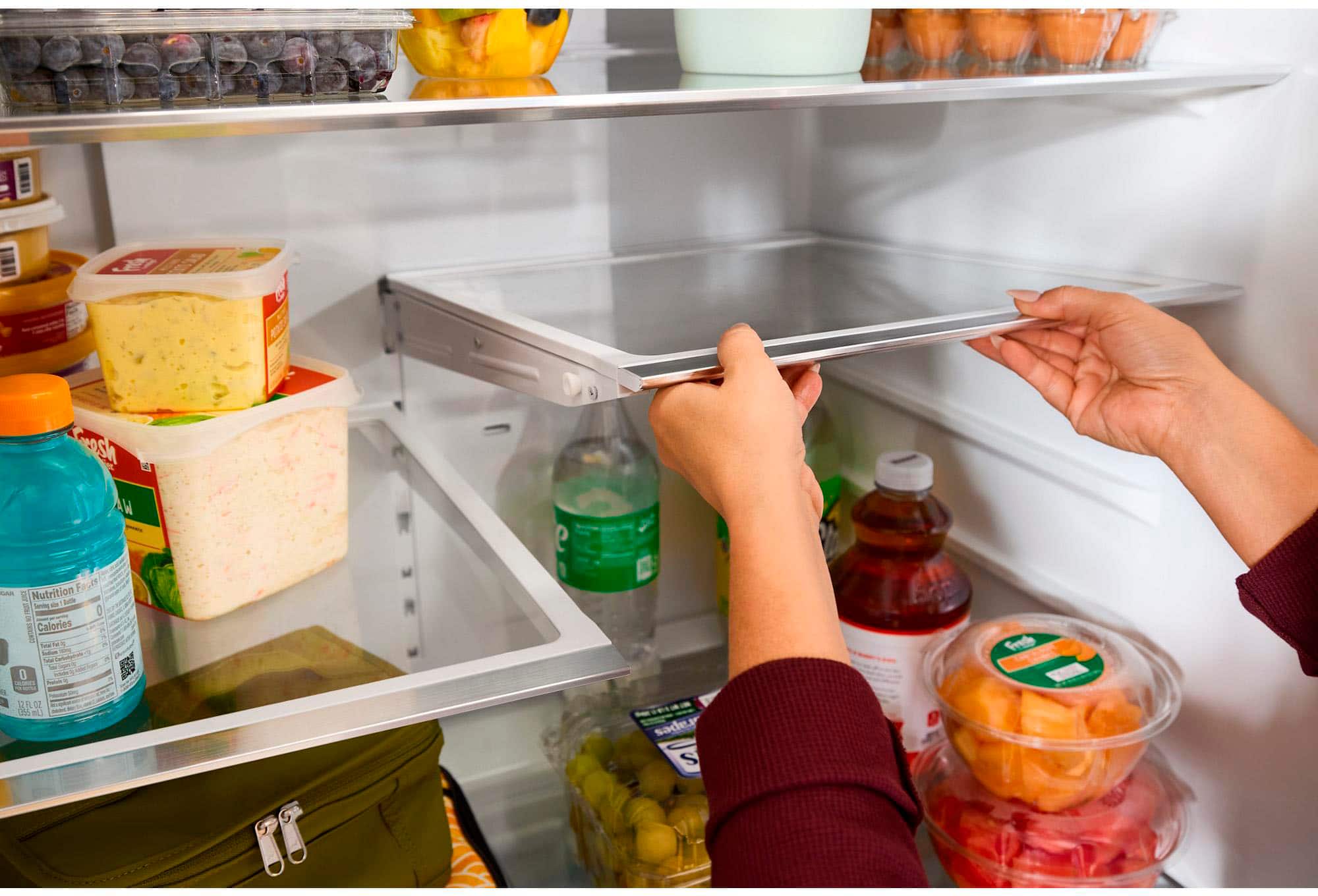 A person is standing in front of an open refrigerator, holding a bottle of water. The refrigerator is filled with various food items, including a bowl of fruit and a carton of yogurt. The person is likely looking for something to eat or drink.