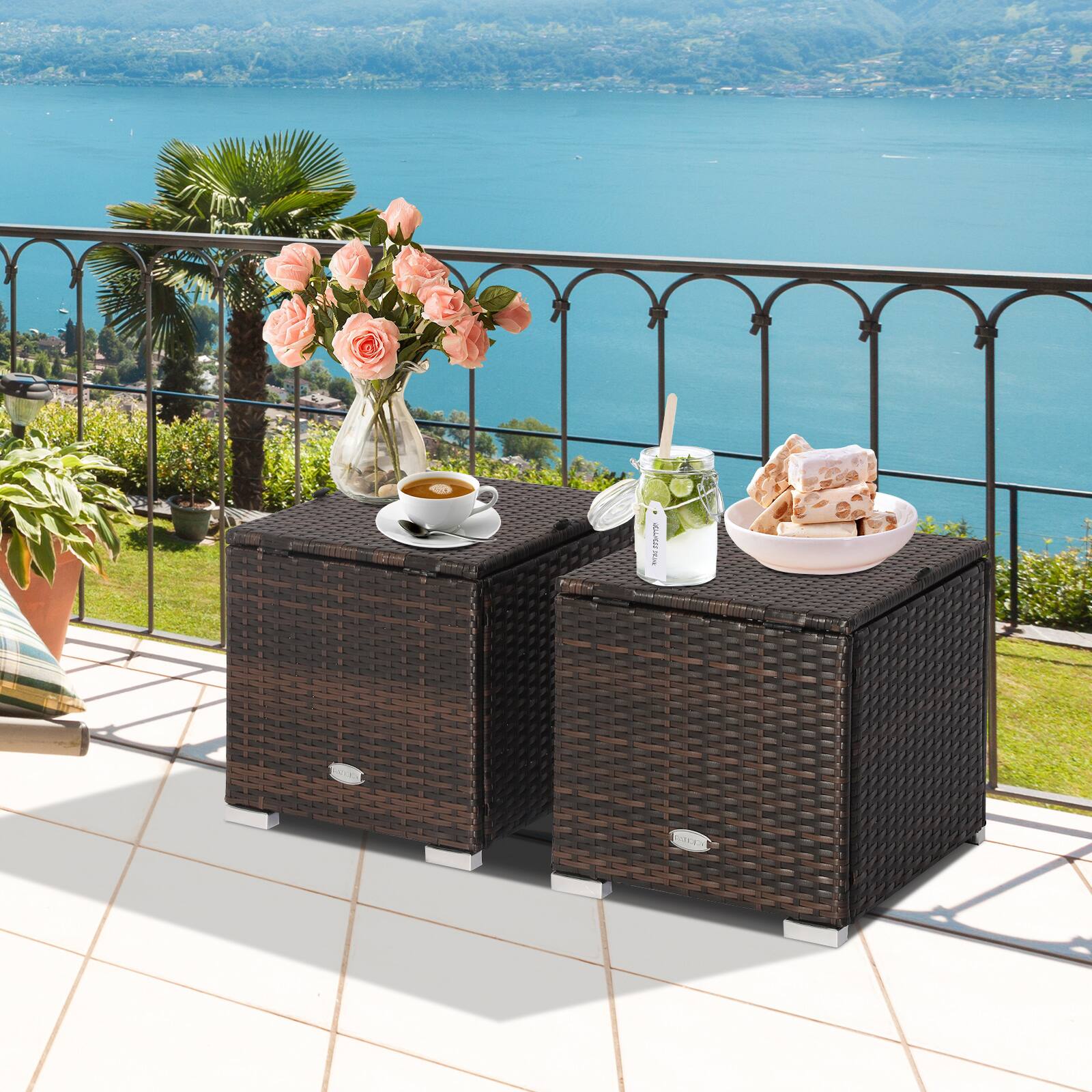 The image features a patio with a table and two wicker chests on it. The table is topped with various items, including a cup of coffee, a bowl of food, a vase with flowers, a bottle, and a spoon. The chests are placed on the table, and there is a chair nearby. The scene is set by a beautiful view of the water, making it an ideal spot for a relaxing meal or a casual gathering.