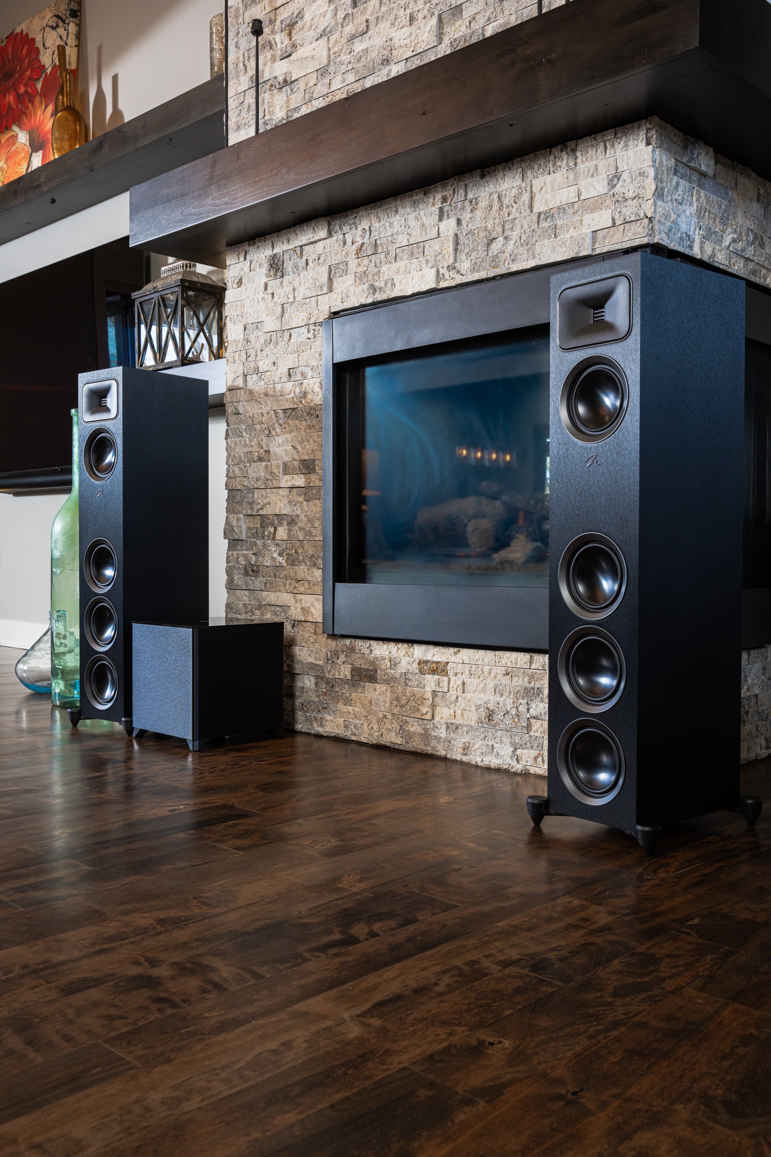 The image shows a living room with a stone fireplace. There are two large speakers on either side of the fireplace, creating a home theater system. The room also features a wooden floor and a TV mounted on the wall. The scene is set in a well-decorated living area, with a variety of decorative elements such as vases, bottles, and a clock. The overall atmosphere of the room is cozy and inviting, perfect for relaxing and enjoying some entertainment.