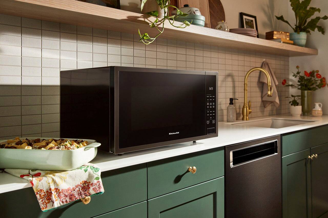 The kitchen features a black microwave sitting on top of a counter. The counter is green and has a towel hanging from it. The kitchen also has a sink and a potted plant nearby.
