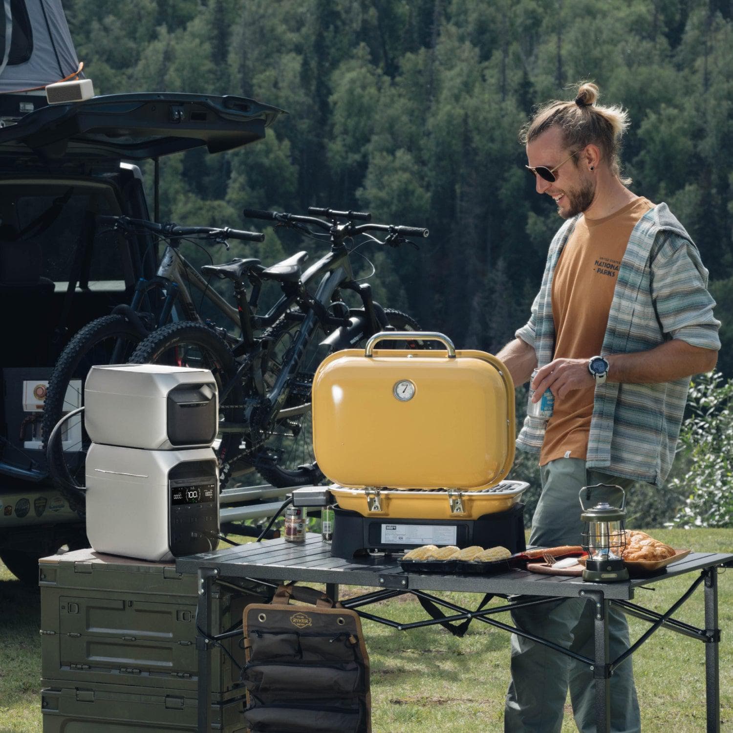 The image shows a man standing next to a yellow portable grill, which is placed on a table. The table also has a cooler and a microwave on it. The man is smiling and appears to be enjoying his time outdoors. There are two bicycles in the background, and a truck can be seen nearby. The scene suggests that the man is preparing food for a picnic or an outdoor gathering.