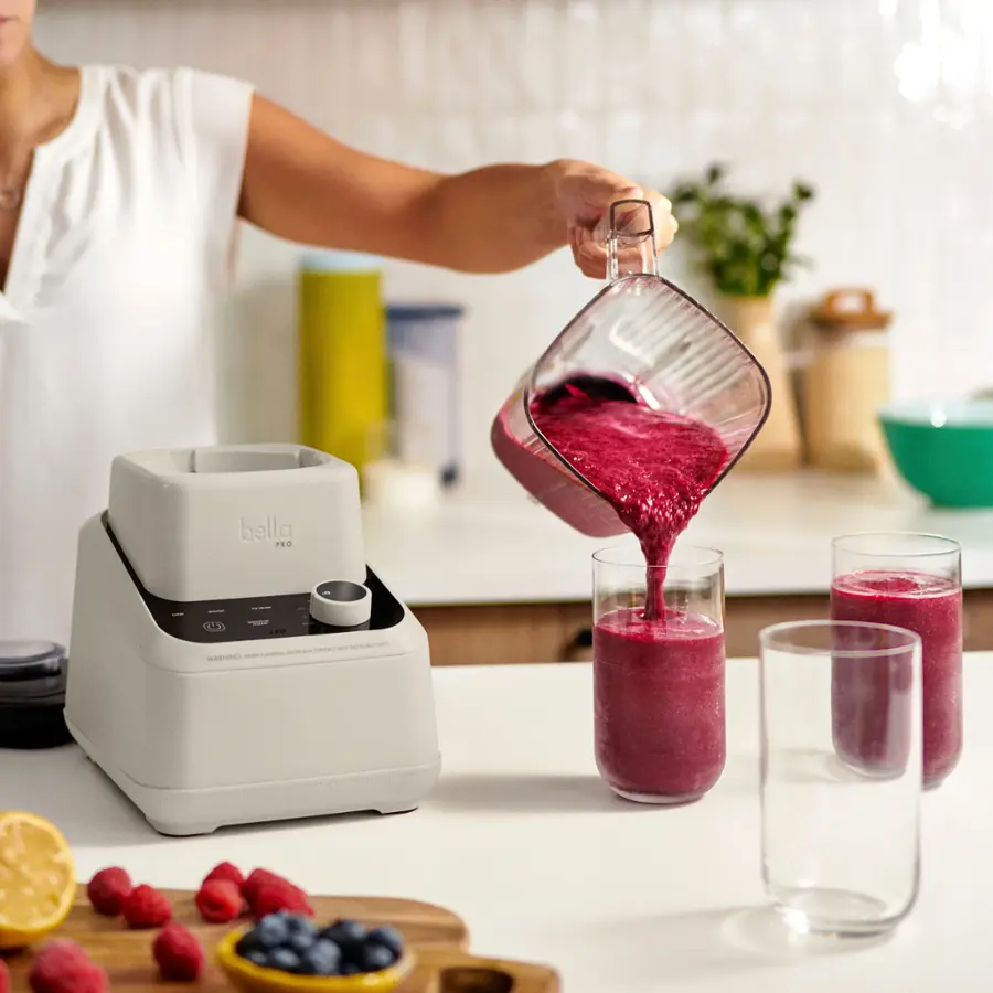 The image shows a woman pouring a drink into a glass using a blender. The blender is placed on a counter, and there are two cups nearby. The woman is wearing a white shirt and is standing in front of the blender. The scene also includes a bowl and a potted plant in the background.