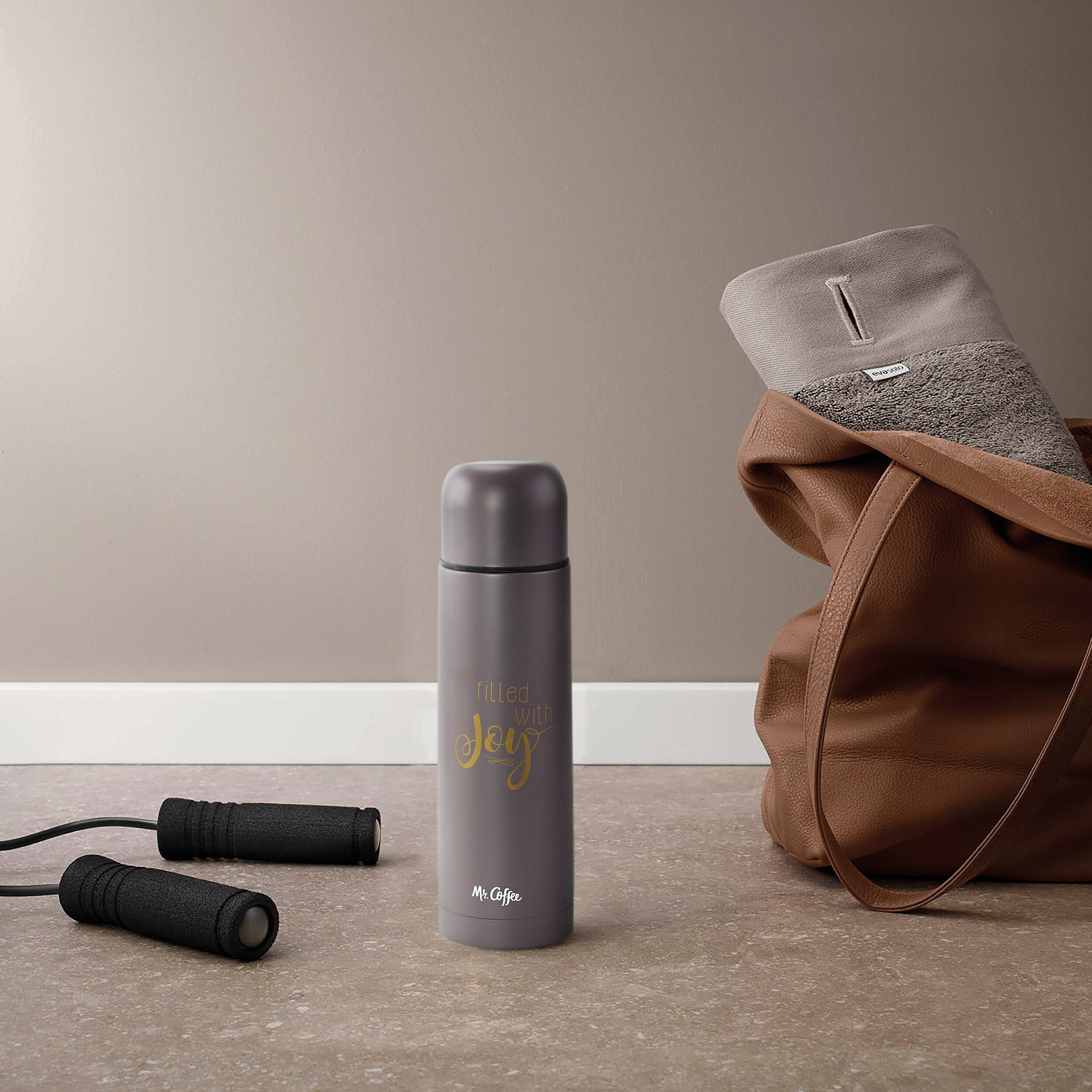 The image features a grey water bottle filled with coffee, placed on the floor next to a brown bag. There are also two black dumbbells nearby, indicating that the person may be preparing for a workout or exercise session. The combination of the coffee, bag, and dumbbells suggests that the person is likely getting ready for a workout or a busy day ahead.