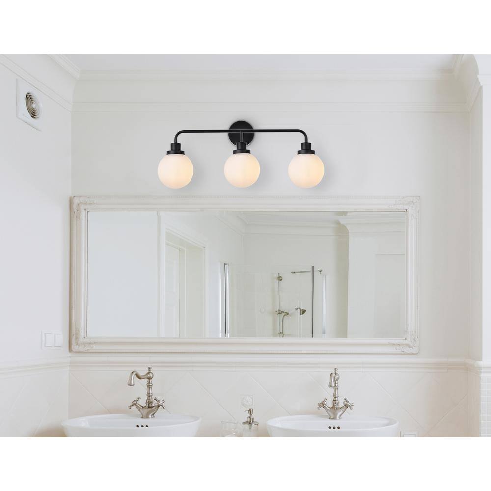 The image shows a bathroom with a large mirror above two white sinks. The mirror has a decorative frame, and there is a black wall-mounted light fixture with three round bulbs above it. The faucets are silver and have a classic design. The walls are painted white, and there is a white tiled backsplash.