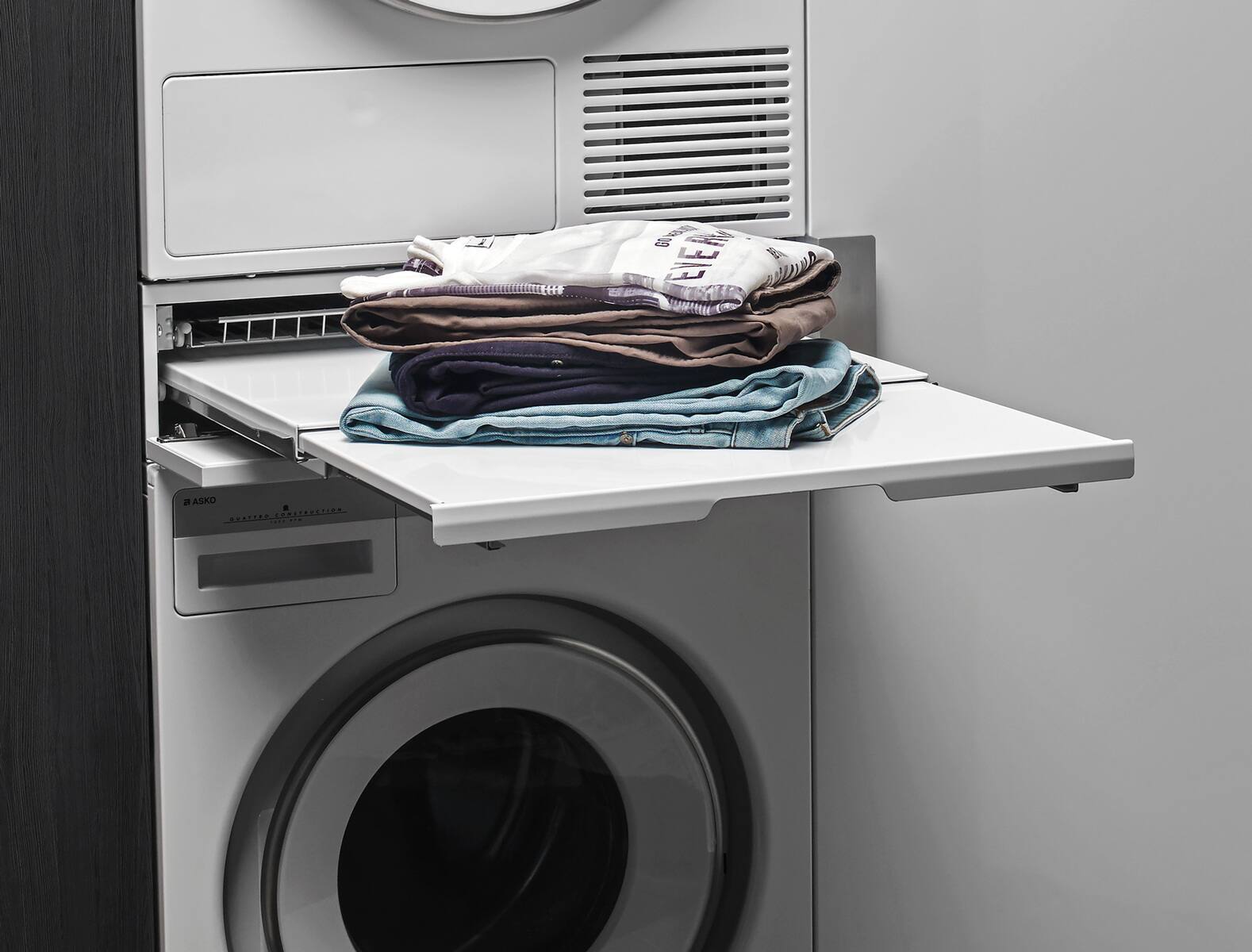 A white washing machine with a shelf on top of it holding a pile of clothes.