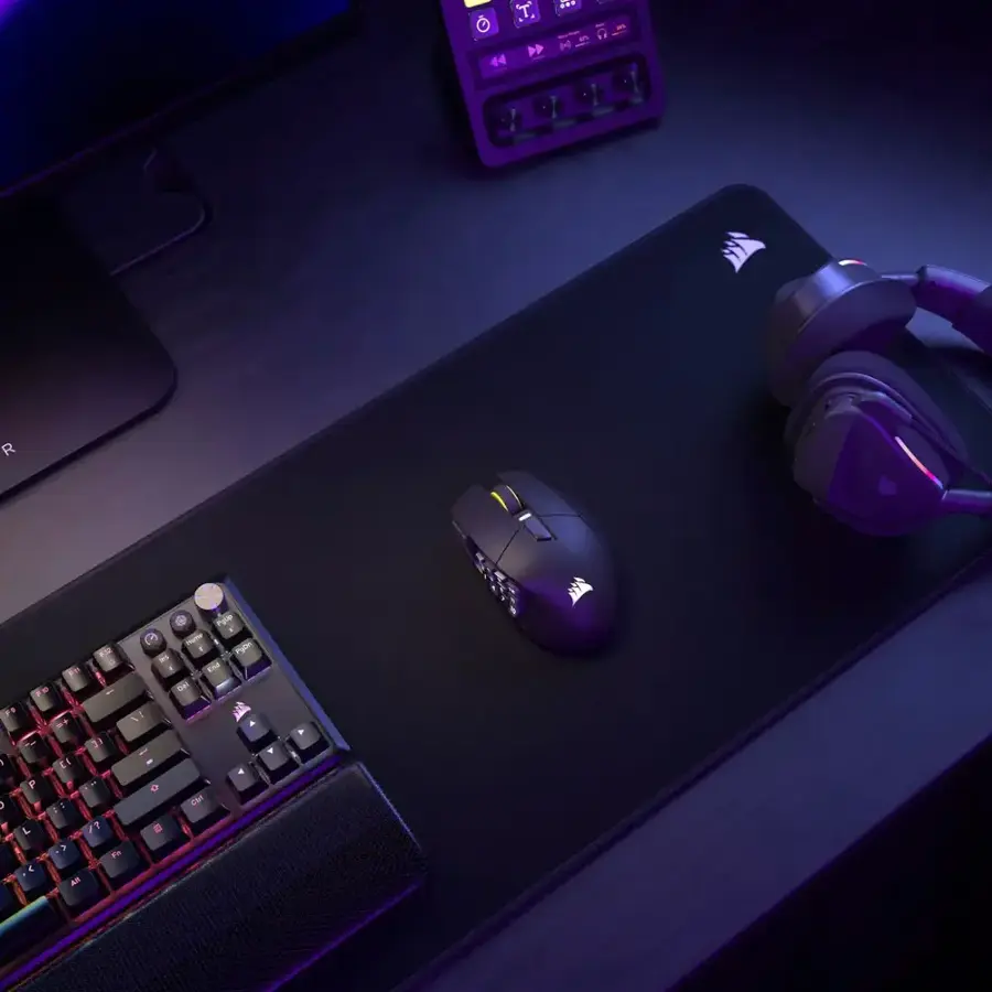 The image shows a computer desk with a keyboard and mouse on a black mouse pad. The keyboard has a purple and black color scheme, and the mouse is black. The desk is set up for a gaming session, with the mouse and keyboard ready to be used.