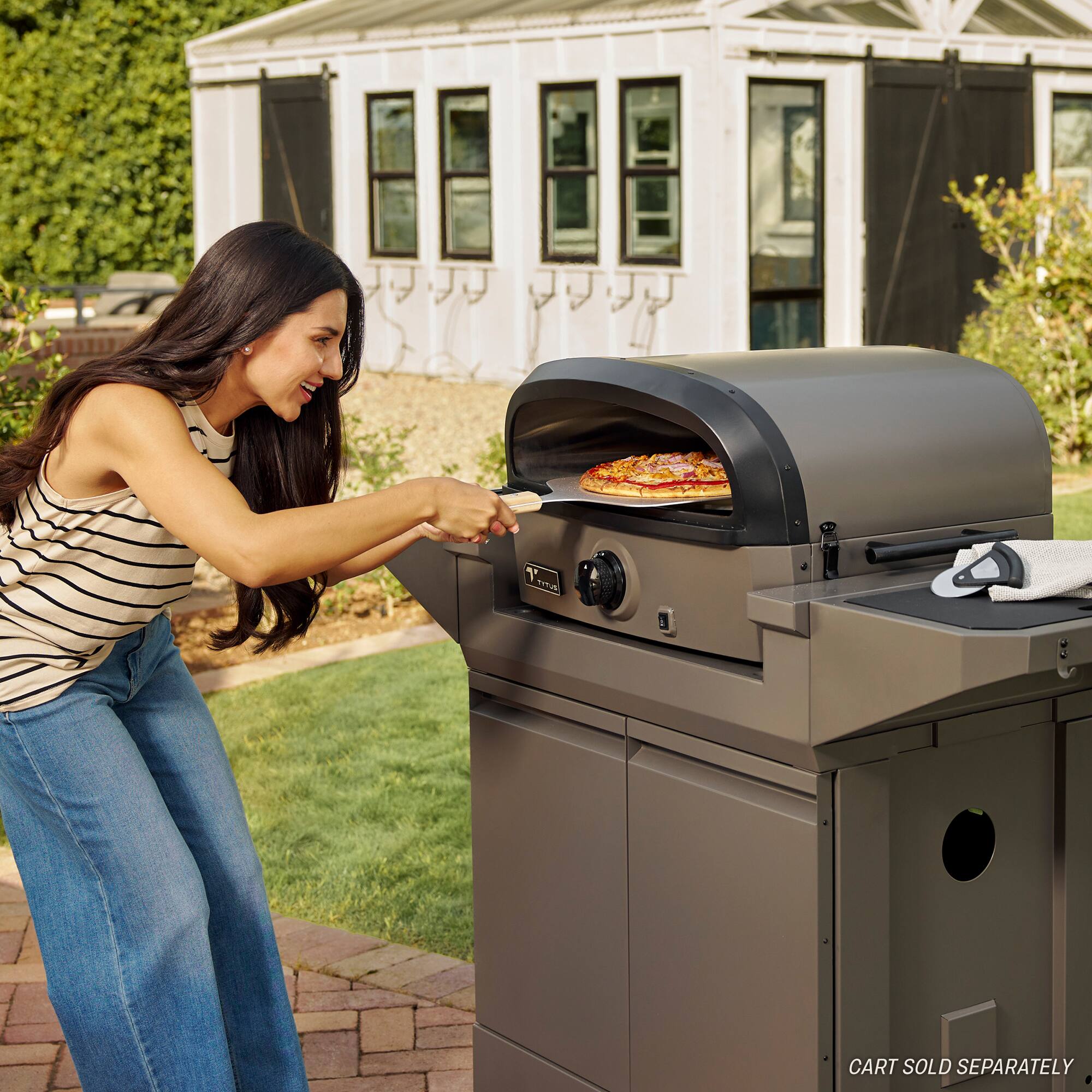The image shows a woman standing in a backyard, placing a pizza into an outdoor grill. The pizza is on a pan, and the woman is using a pizza peel to slide it into the grill. The scene suggests that she is preparing a meal outdoors, possibly for a gathering or a casual outdoor event. The outdoor grill is a key element of this scene, as it provides a unique and enjoyable way to cook food, especially during warm weather.