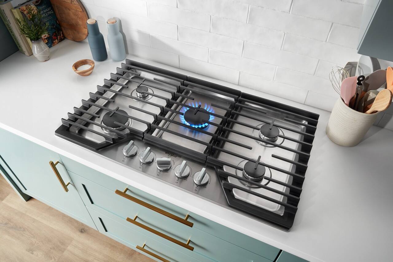 The image features a stove top oven with a blue flame on the burner. The stove is situated in a kitchen with a white countertop and a white wall. There are several bottles placed around the kitchen, with one near the stove, another on the countertop, and a third one further away. Additionally, there are two knives and a spoon located near the stove, possibly for cooking or preparing a meal.