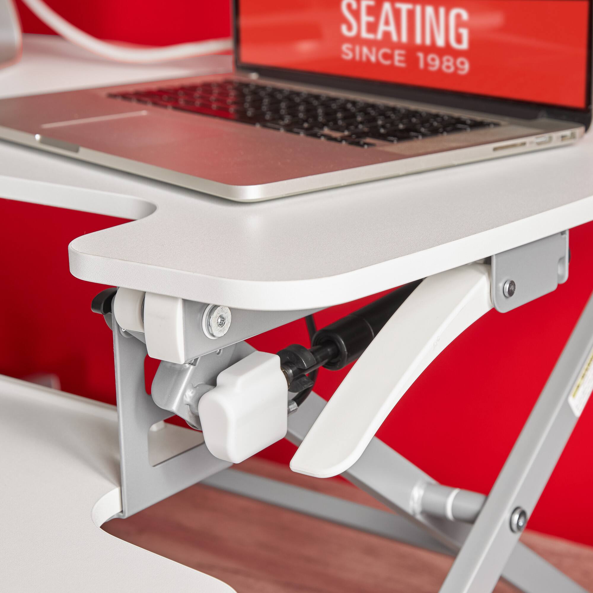 The image features a laptop computer sitting on a white desk, with a red wall in the background. The laptop is positioned on a stand, and there is a red sign above it. The sign reads "seating since 1989," which suggests that the company has been providing seating solutions for over 30 years. The laptop is open, and the desk appears to be well-organized and functional.