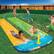 The image shows a group of children playing on a water slide in a backyard. There are at least six children visible, with some of them playing in the water and others standing nearby. The children are enjoying themselves and having fun on the slide.
