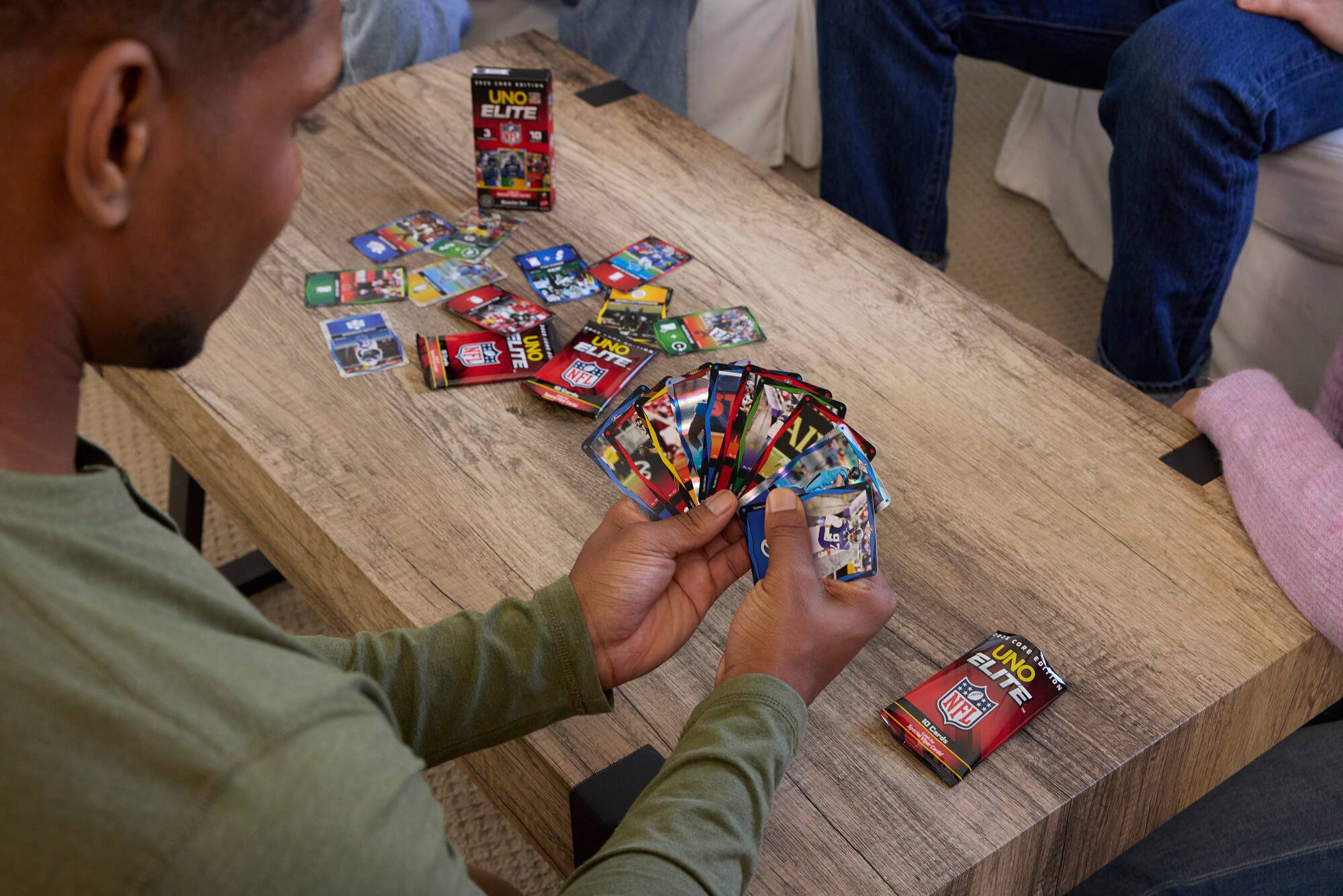 The image features a group of people sitting around a wooden table, playing a card game. The game they are playing is called "UNO Elite." The table is covered with a variety of cards, including NFL cards, which are part of the game. There are at least four people visible in the scene, with one person holding a card in their hand. The table is surrounded by chairs, and the people are engaged in a fun and social activity.