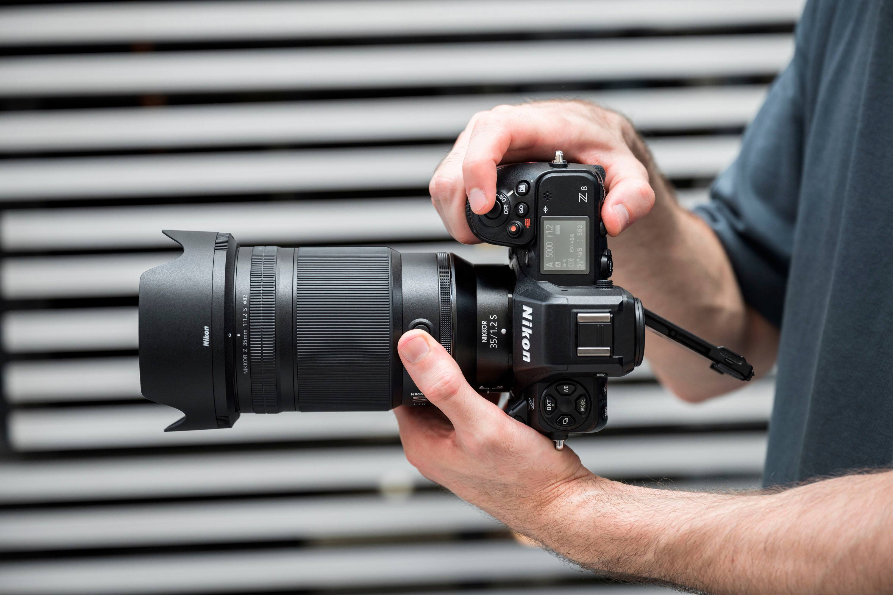 The text on the image reads: "Nikon 8 u 1:1.2 35mm - NIKKOR - NO OFF S NIKKOR 35/1.2  do Nikon N 8 Z8  F1.2 562 5000 100." This text is likely a description or specifications for the camera lens being held by the person in the image.