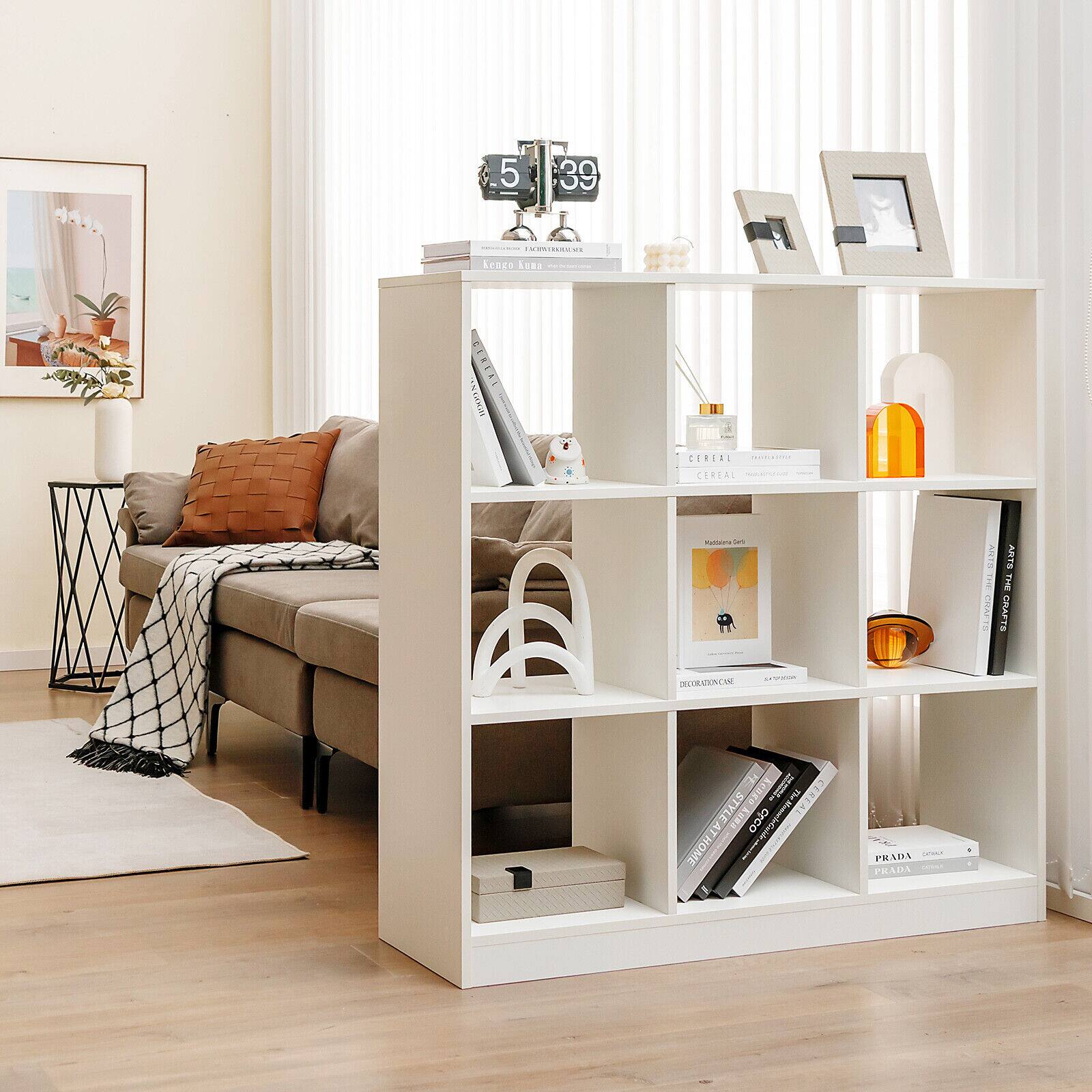 A white bookshelf with a variety of books and items on it. The books are arranged in different sizes and orientations. The bookshelf is located in a living room, and there is a couch nearby. The room also features a clock and a vase, adding to the cozy atmosphere.