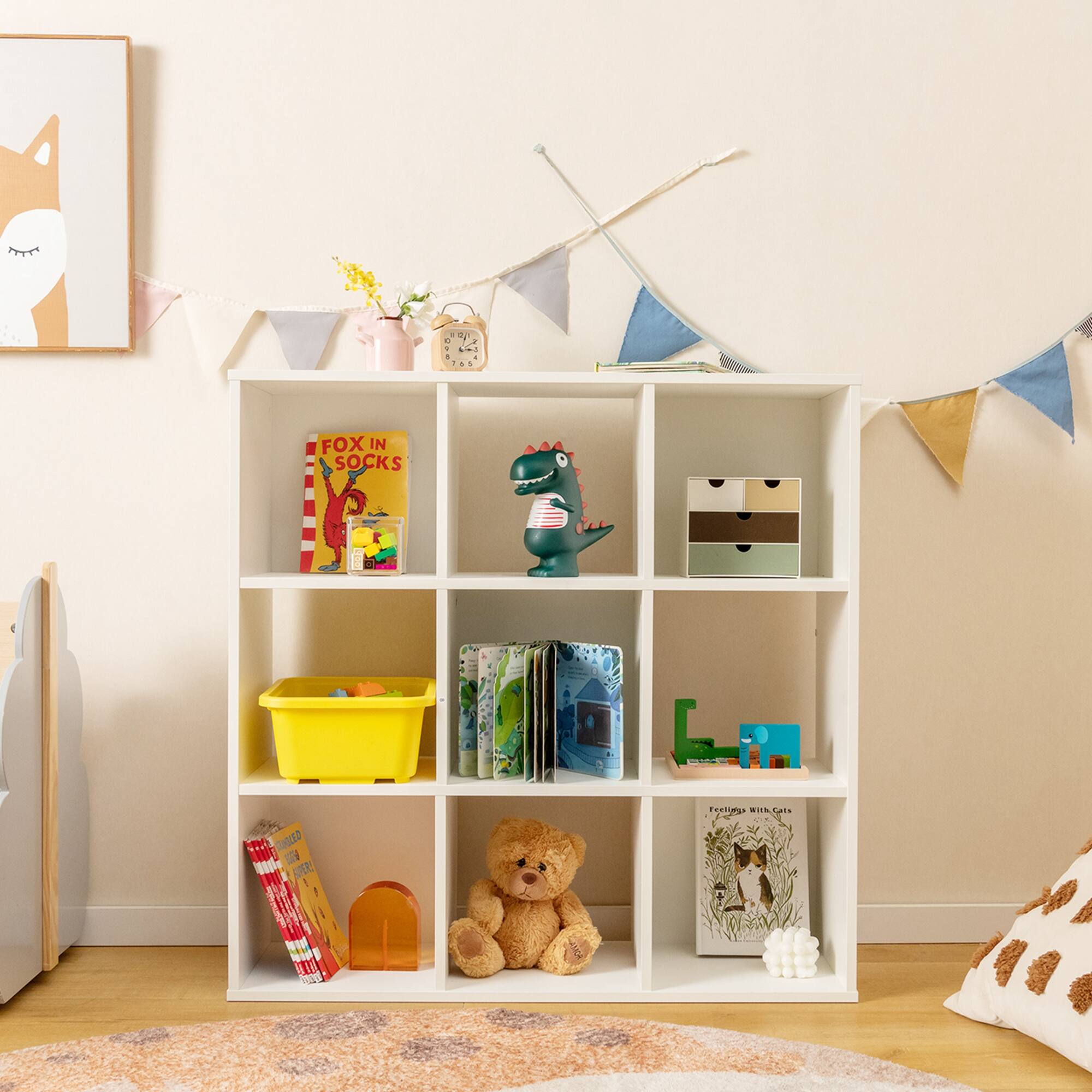 The image shows a white bookshelf filled with various books and toys. There are several books of different sizes and colors, as well as a teddy bear sitting on the shelf. The bookshelf is located in a child's room, and there is a painting of a fox on the wall. The scene also includes a chair and a couch, creating a cozy and inviting atmosphere for the child to read and play.