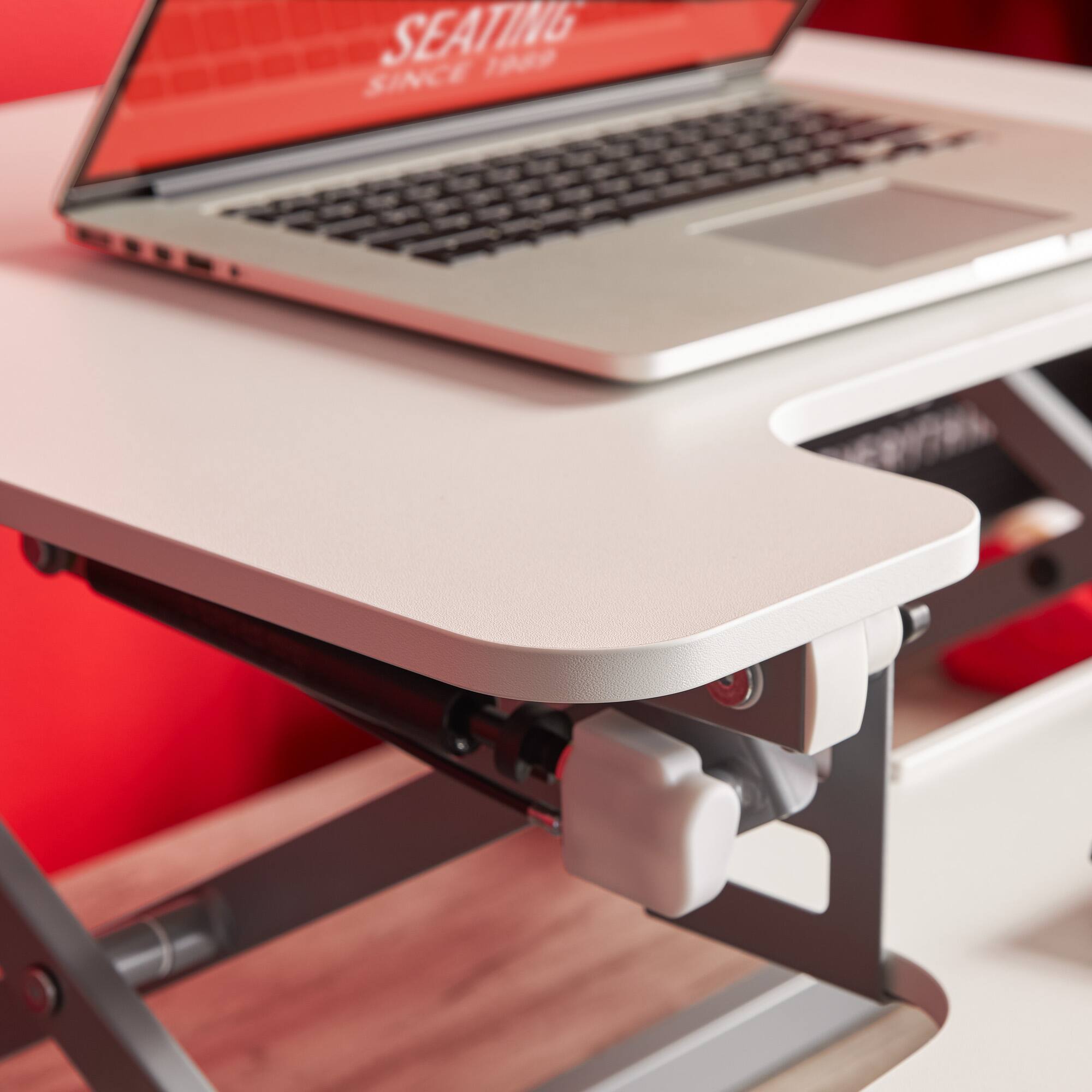 The image features a laptop computer sitting on a desk. The laptop is positioned on a stand, which elevates it to a more ergonomic height. The desk is white, providing a clean and minimalist backdrop for the laptop. The laptop is open, and the keyboard is visible, indicating that it is ready for use. The overall scene suggests a comfortable and functional workspace.